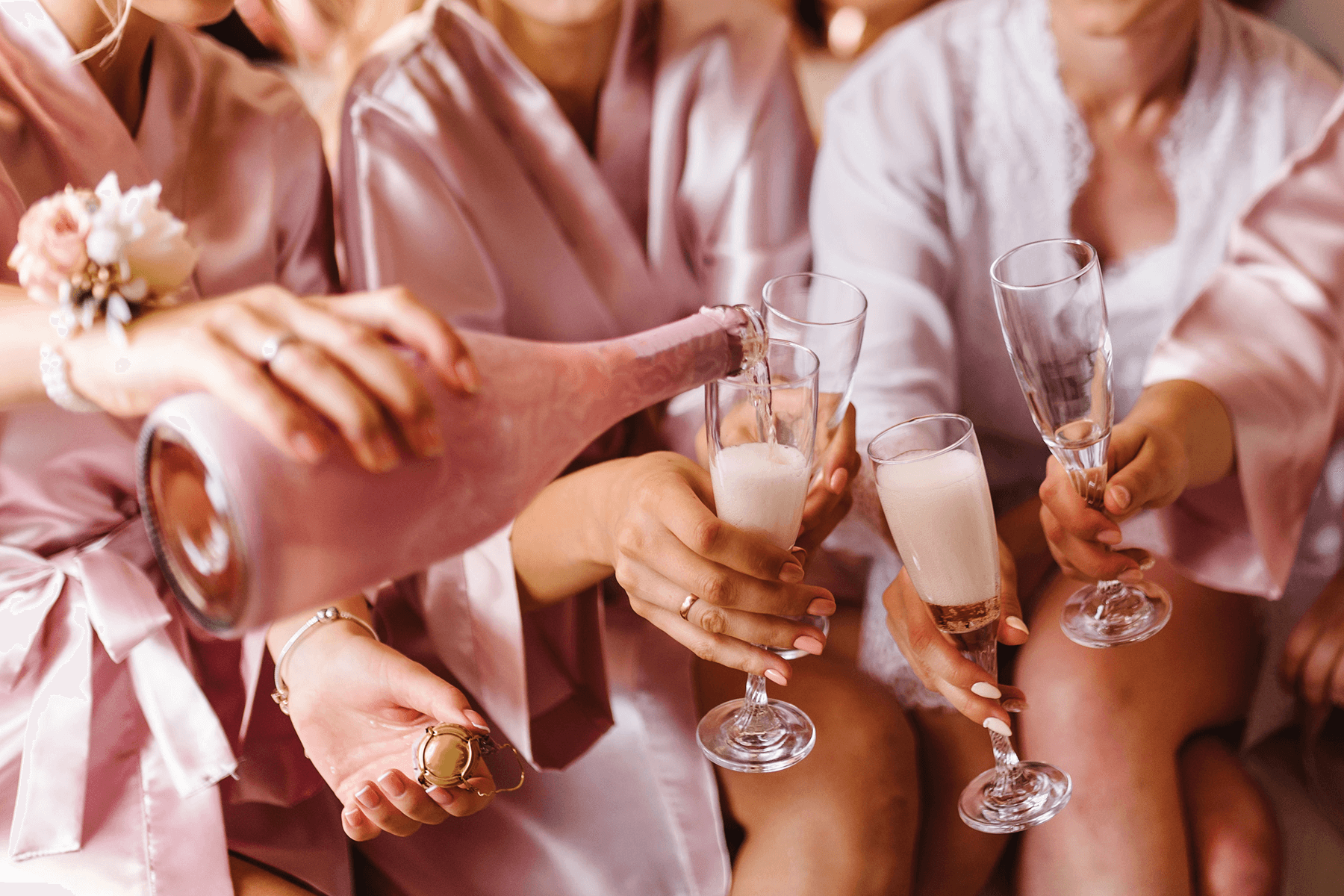 Three women in satin pink robes pour a pink bottle of Champagne into Champagne flutes.