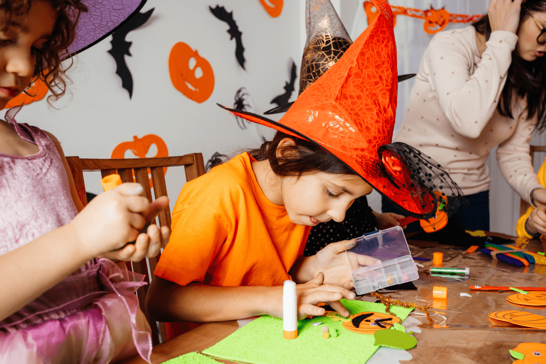 A child in an orange witch hat and orange shirt glues a felt jack-o'-lantern onto a green felt sheet at a wooden craft table, next to a child in a purple witch hat and pink dress, with an adult and more children in the background in front of a wall decorated with paper pumpkins and bats.