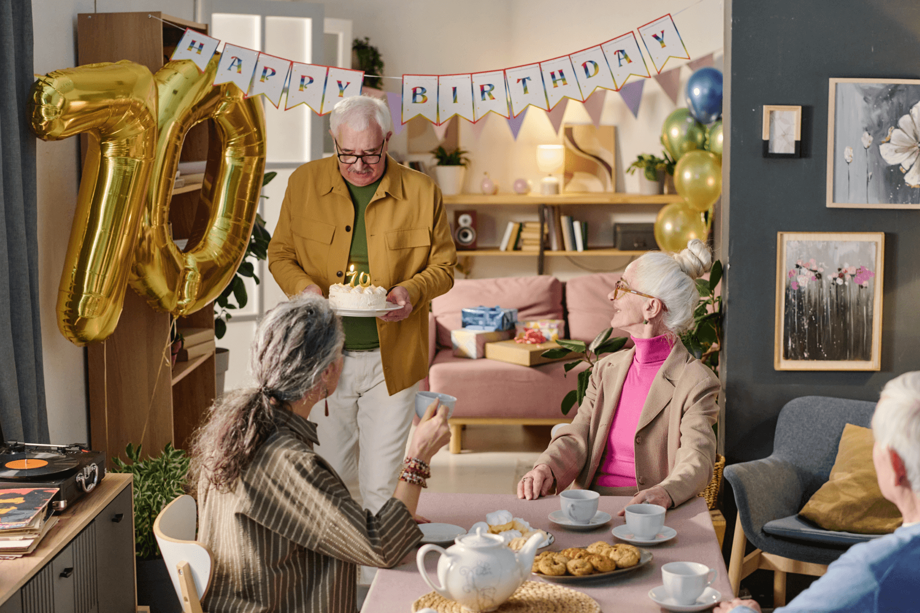 A man holding a birthday cake with lit candles, plus a ‘happy birthday’ garland and ‘70’ gold balloons.