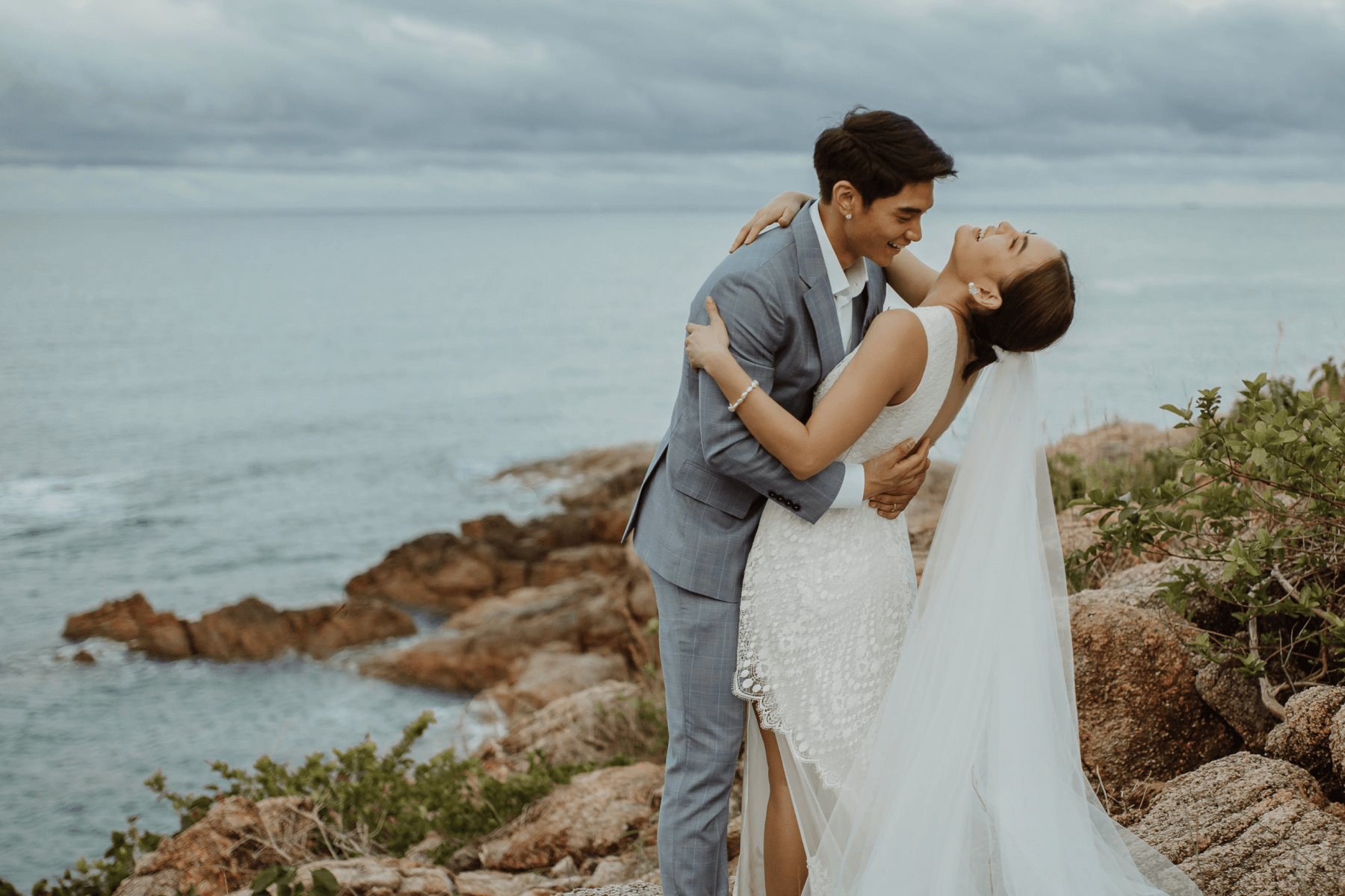 A man in a gray suit embraces a woman in a white wedding gown on a coastline.