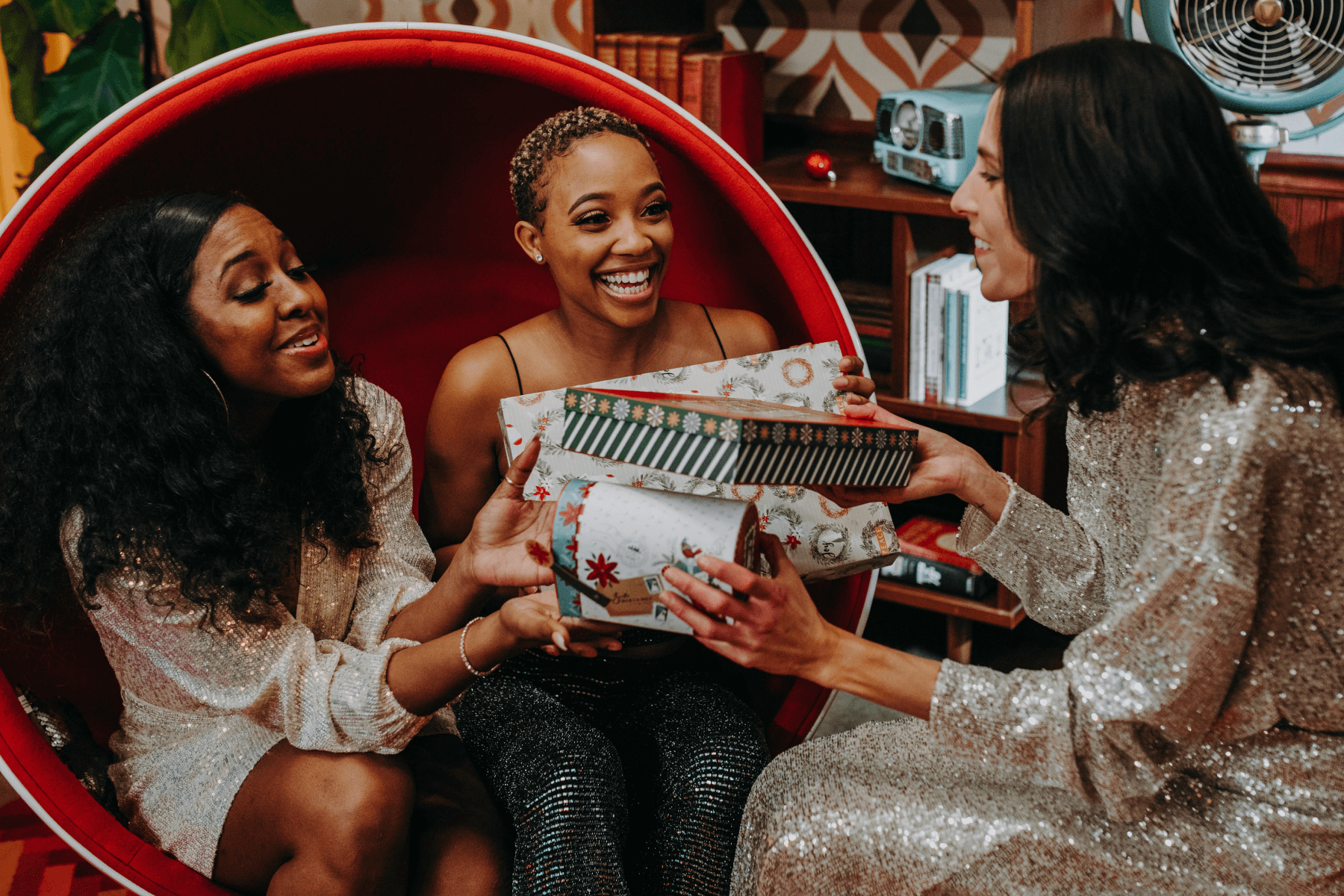 Three women smiling in sparkly outfits holding wrapped Christmas presents.