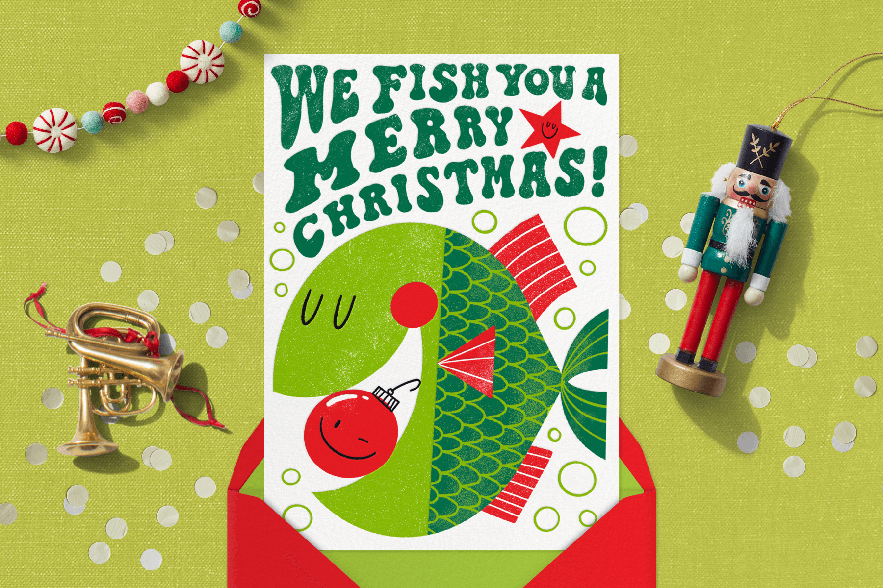 A card with a green fish swallowing a red Christmas ornament and the words “we fish you a Merry Christmas” surrounded by ornaments.