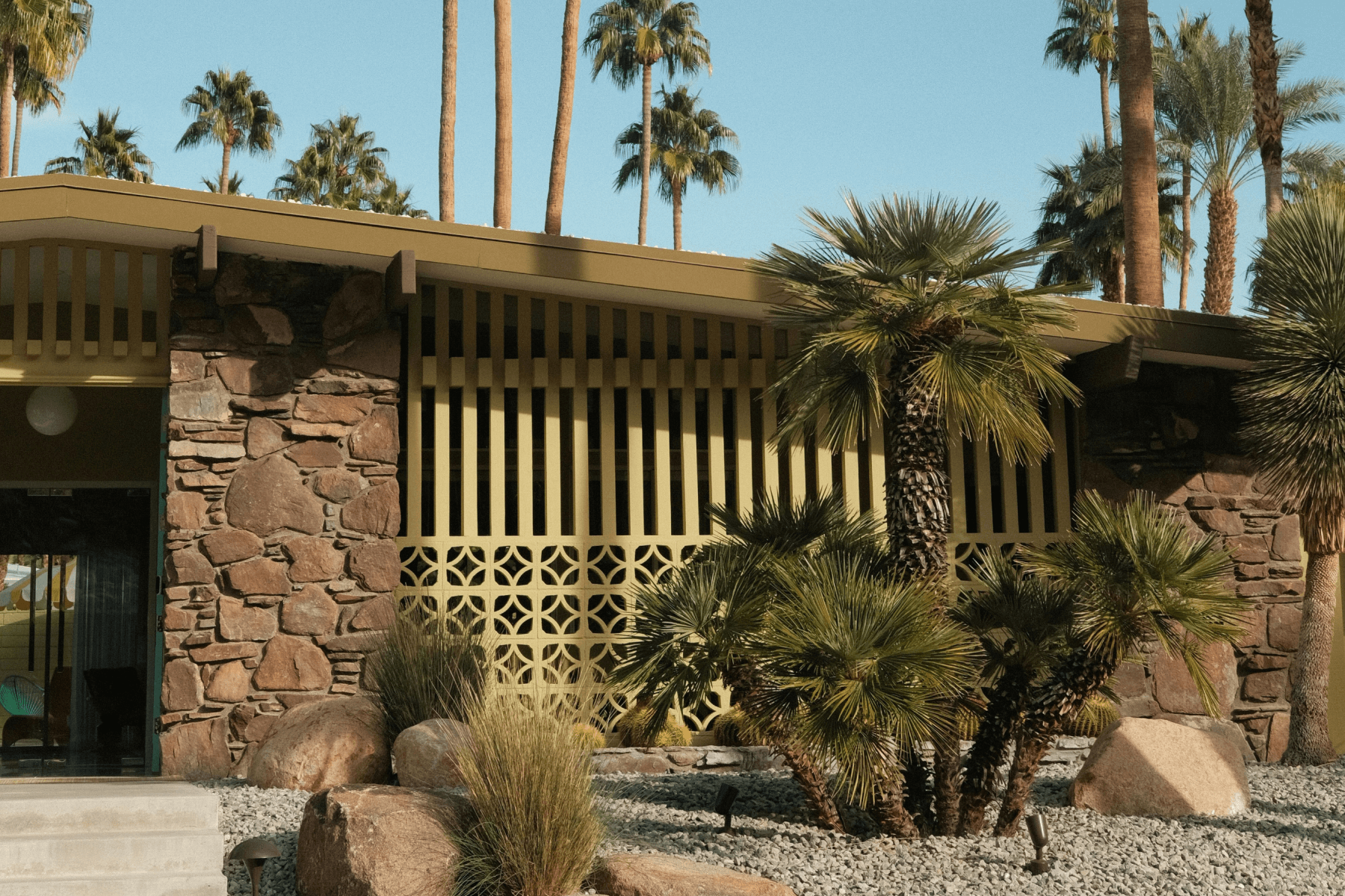 Brown and yellow midcentury-modern house with a rock-filled front yard surrounded by palm trees.