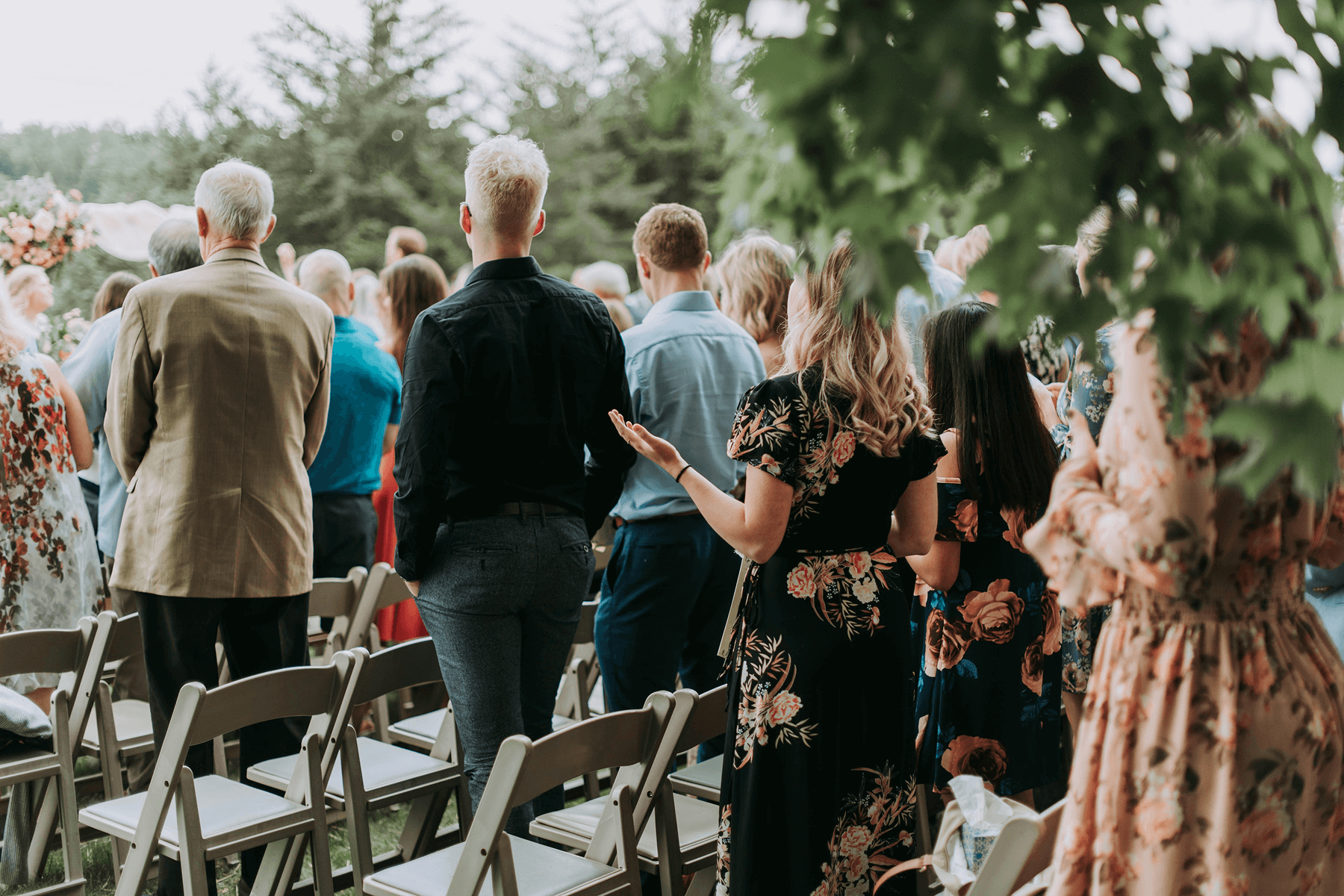 Guests in cocktail attire at an outdoor wedding stand in front of chairs at a ceremony.
