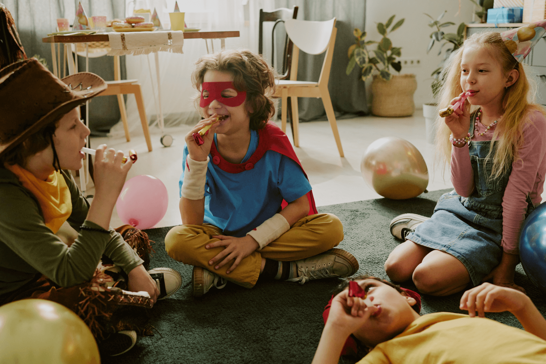 A photograph of four children sitting on a rug wearing party hats and costumes while blowing noisemakers.