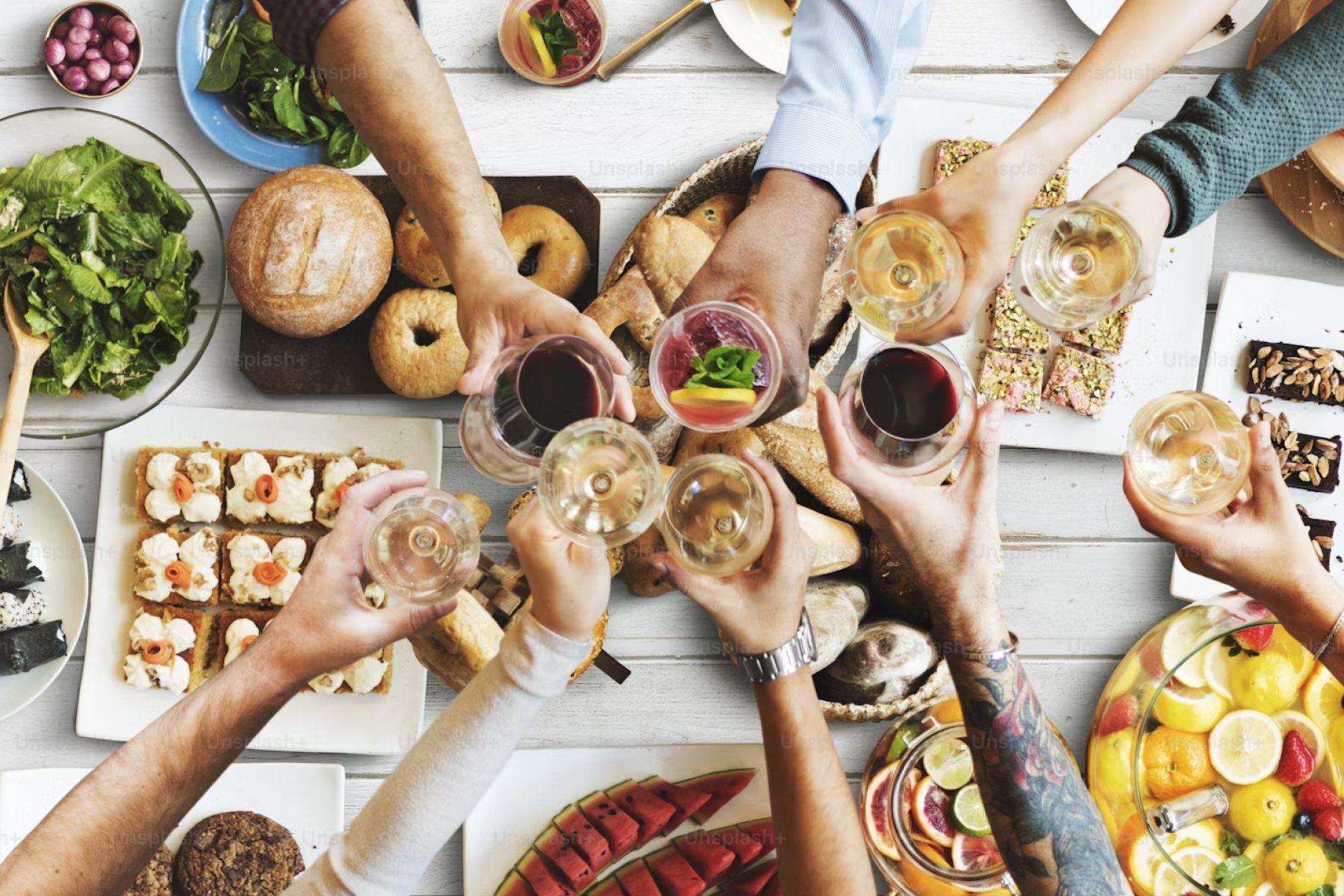 Hands holding drinks and doing cheers over a table full of breakfast foods.