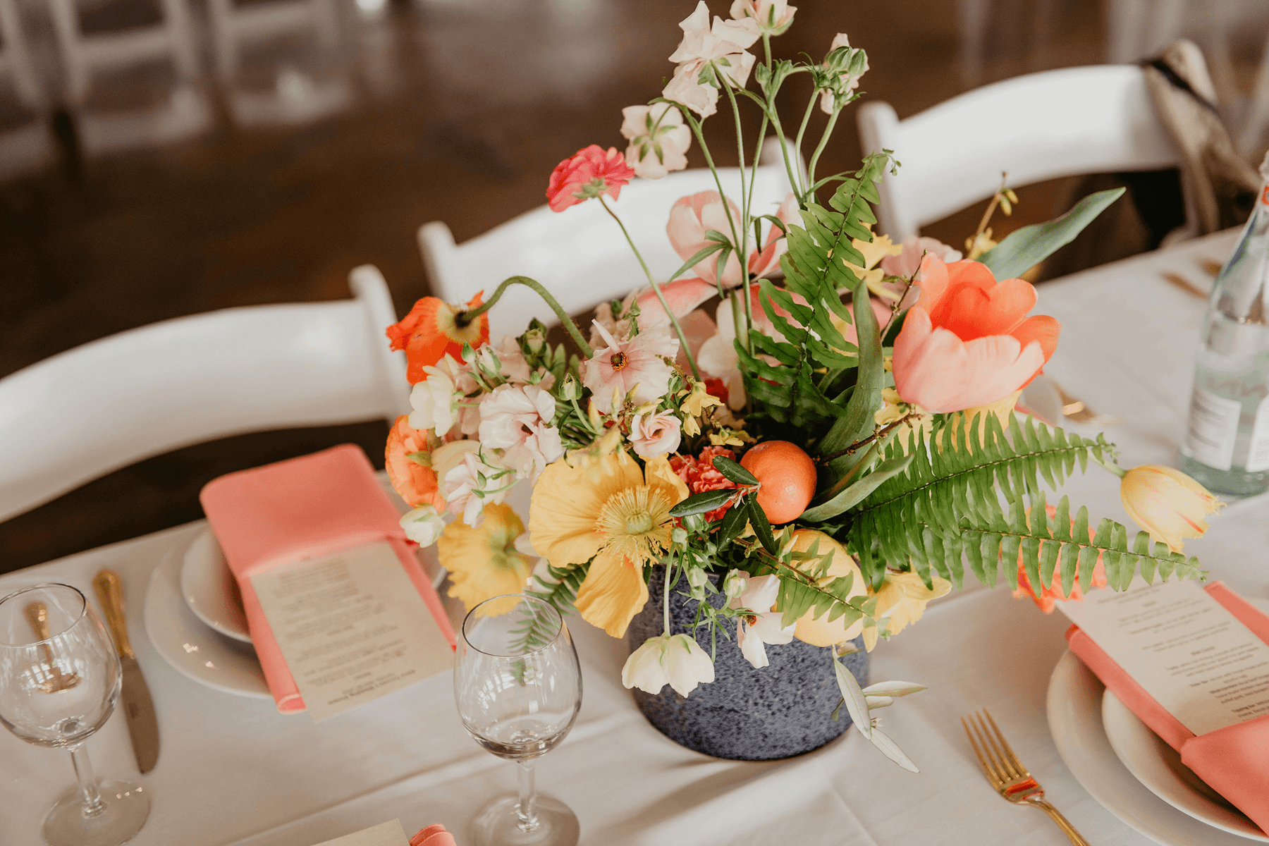 A wedding dinner tablescape featuring a white tablecloth and chairs, peach napkins, gold silverware, and a peach and yellow floral arrangement.
