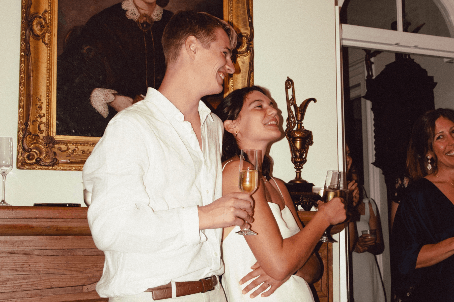 A young man and woman dressed in white holding champagne glasses and hugging.