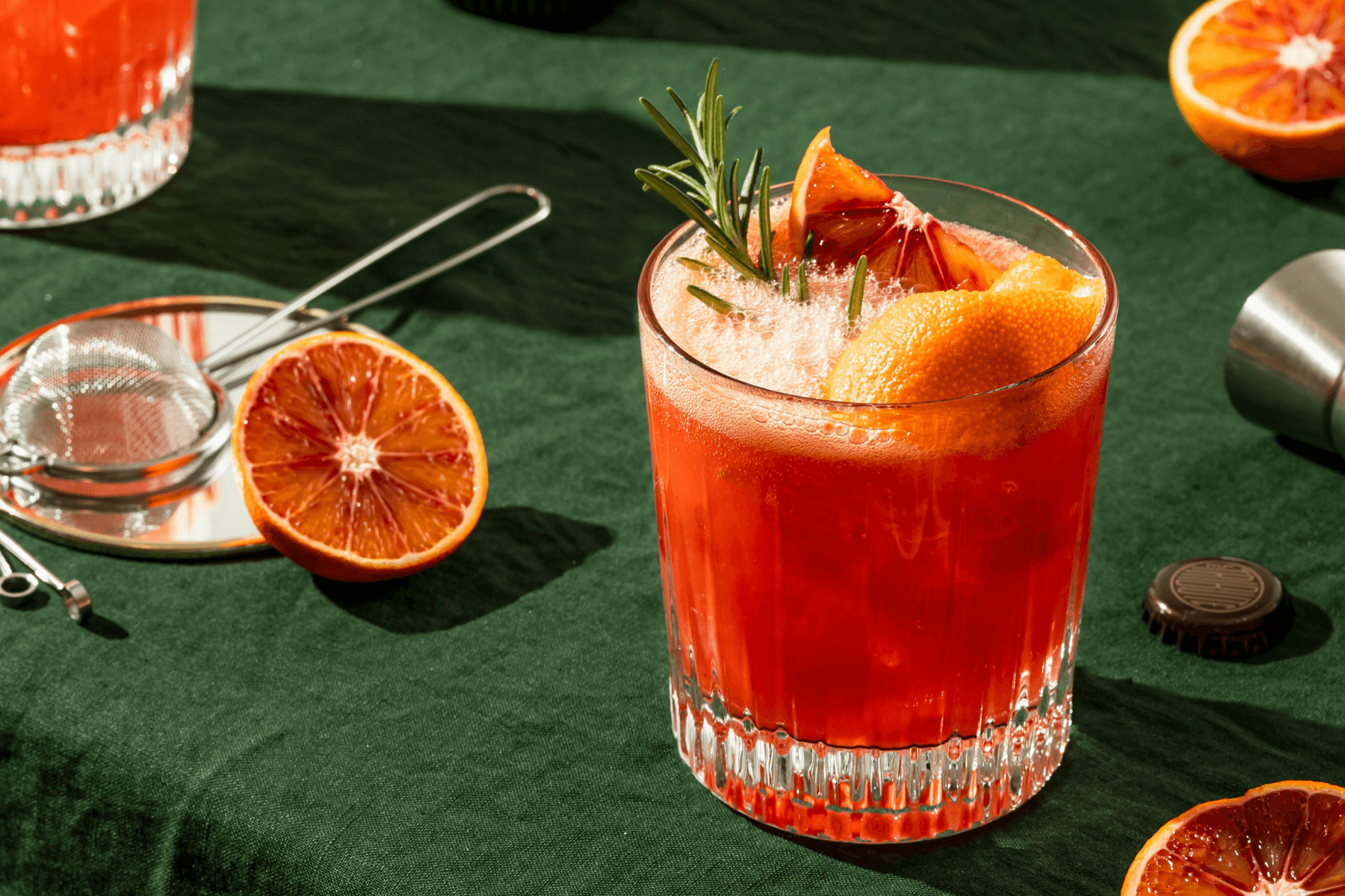 A cocktail glass filled with a red drink, garnished with slices of fruit and a sprig of leaves, on a table with a dark green tablecloth.