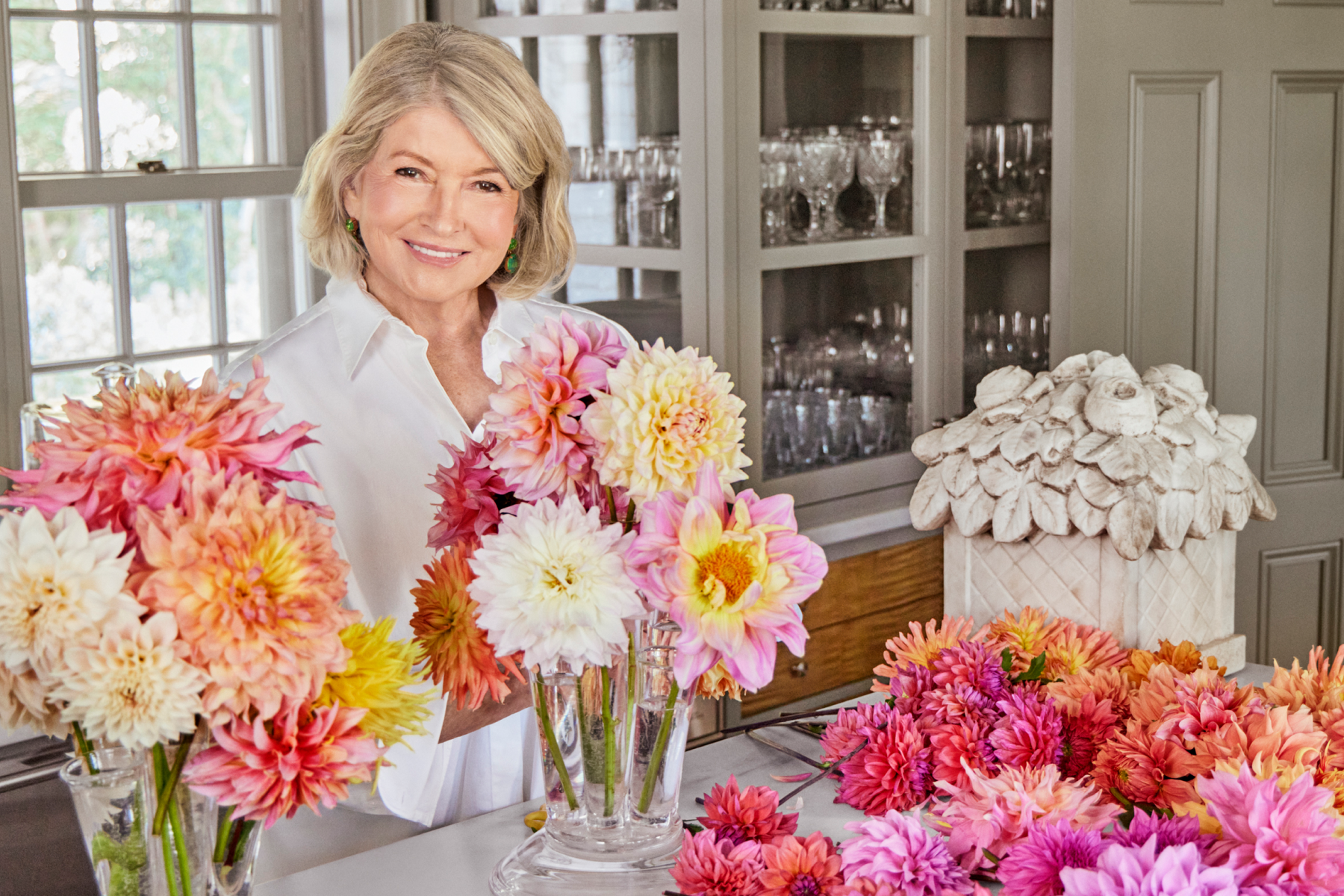 Martha Stewart stands smiling in a kitchen surrounded by pink, white, and orange flowers that she is arranging in clear vases.