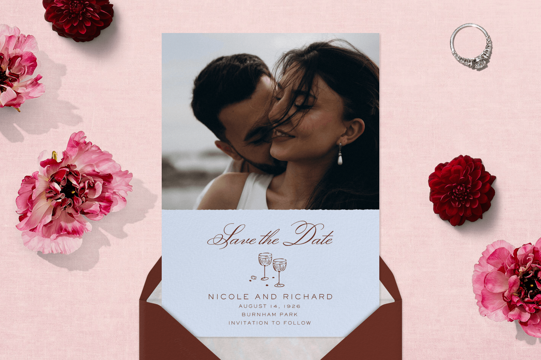 A save the date invitation with an image of a happy couple against a burgundy envelope and a pink textured background with red and pink flowers and an engagement ring prop.