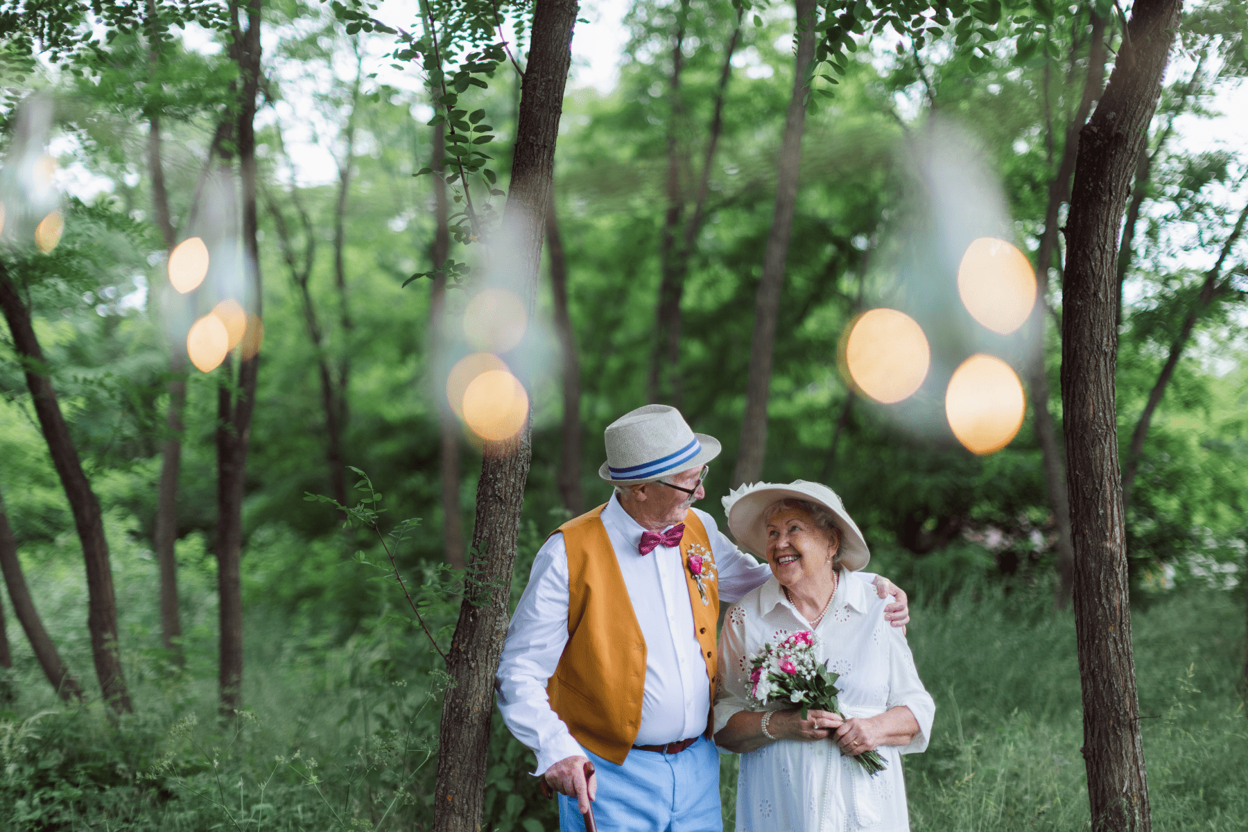 A man and woman dressed in formal wear smiling at each other and hugging under string lights outside.