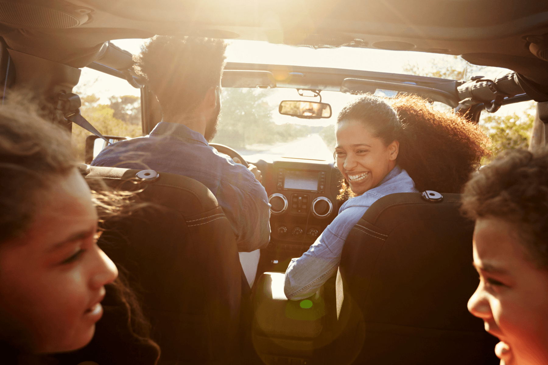 A man, woman, and two children in a car on a sunny day. The woman is smiling and looking back at the two children in the back seat.