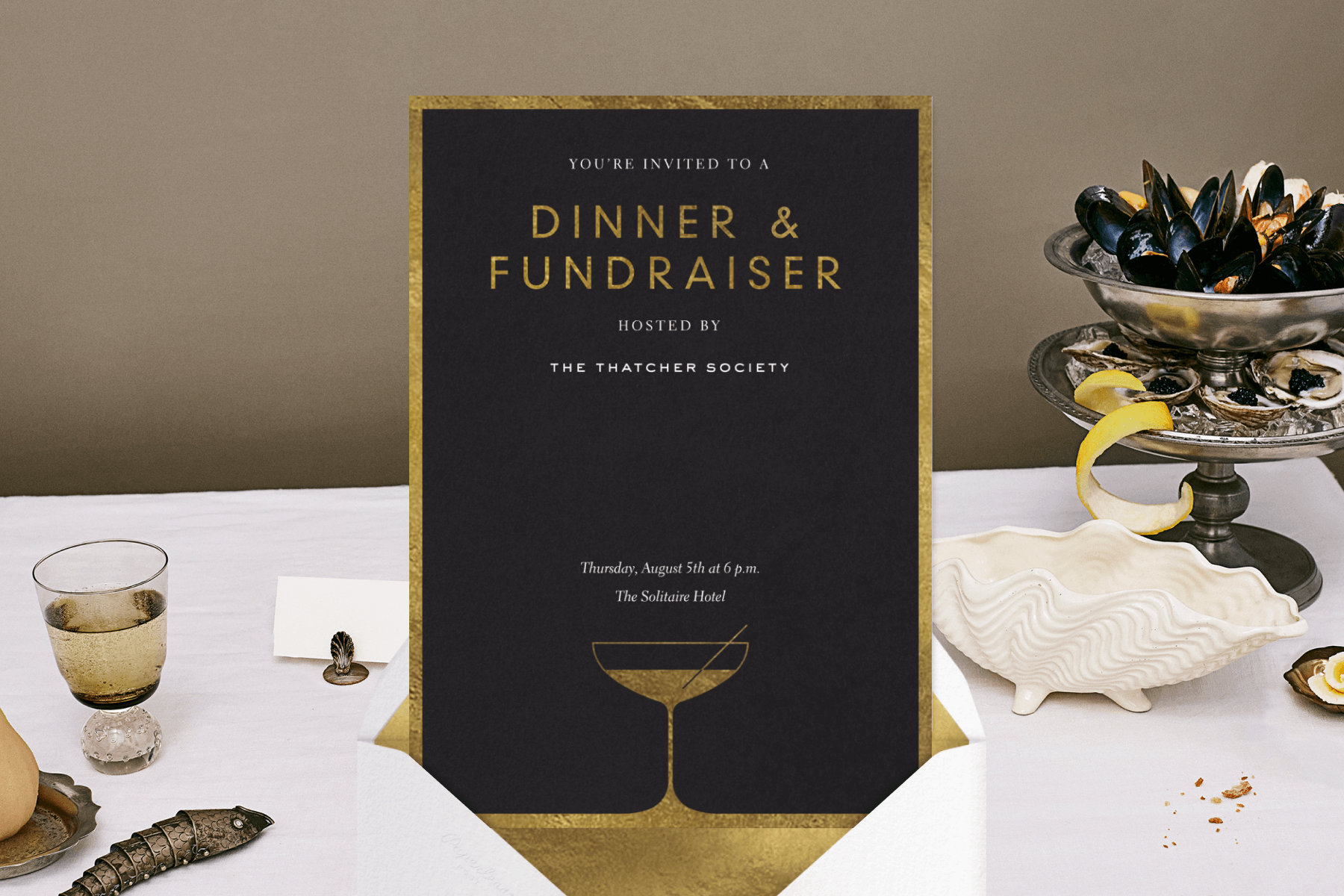 An invitation to a dinner and fundraiser event in gold and black on a white envelope with gold liner, set over a photograph showing a dinner party table with shellfish, drinks, and place cards. 