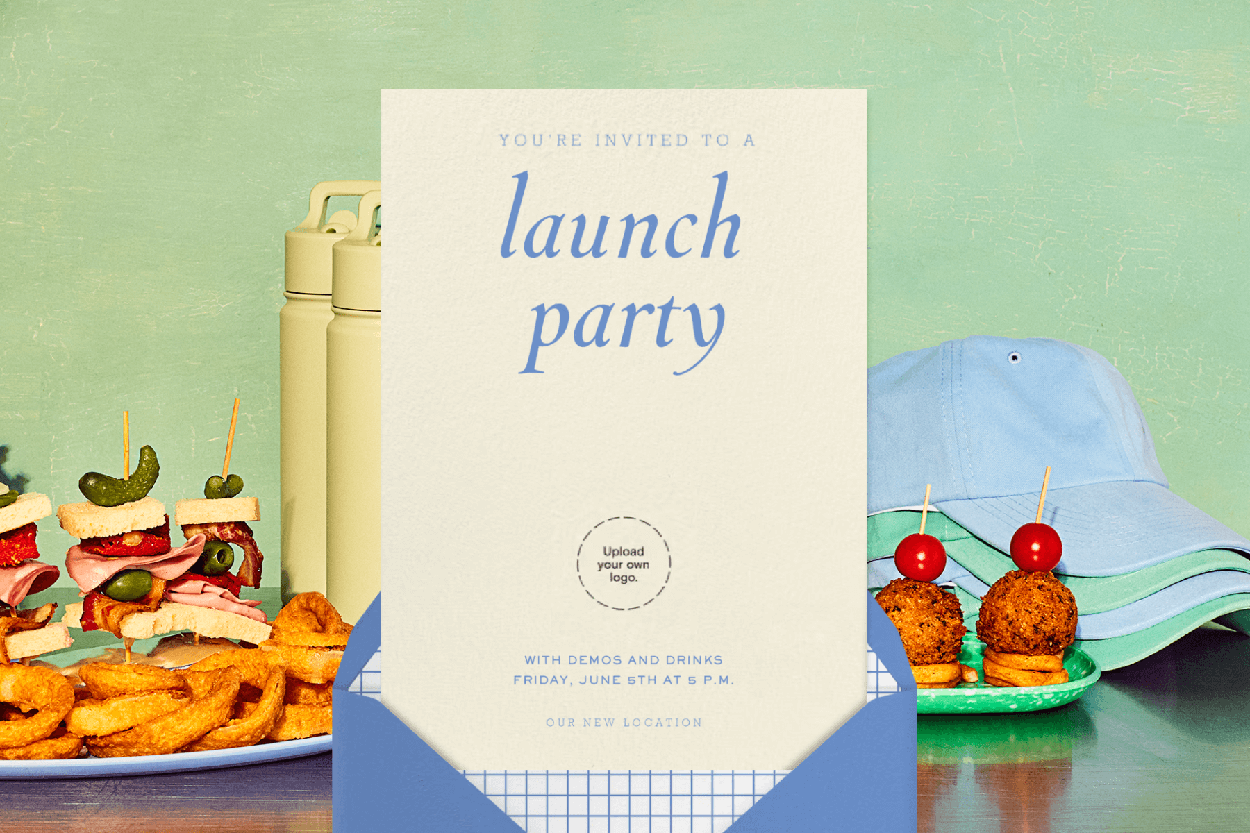 A beige launch party invitation with blue text and a space to upload your own logo in front of a metal table set with plates of food, colorful water bottles, and baseball hats in front of a green wall.