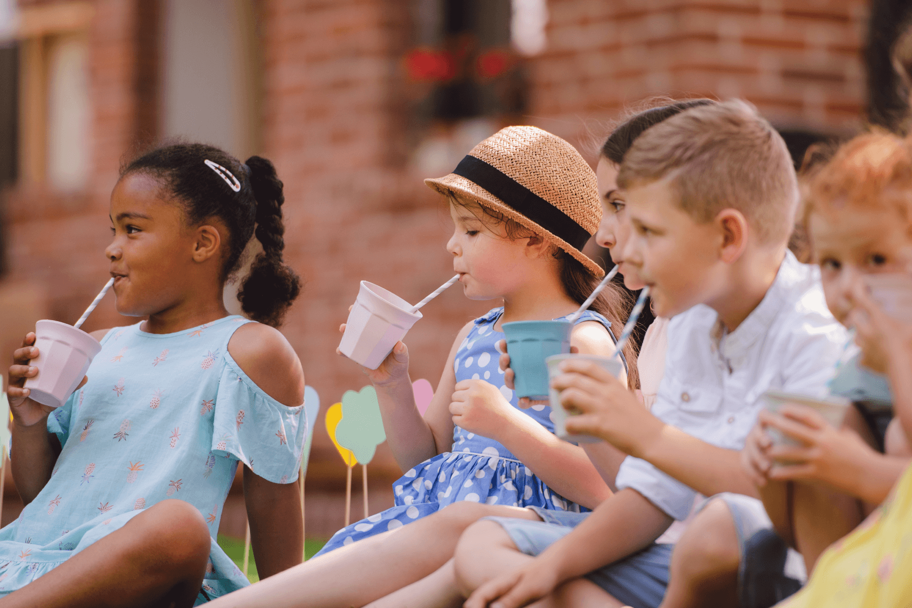 A group of elementary school-aged boys and girls sit on the grass having a picnic and sipping drinks out of cups with straws.