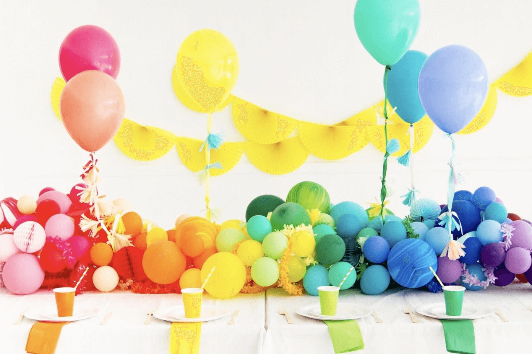 Bunches of rainbow balloons in ROYGBIV order cover a banquet table with colorful party cups and plates in front and several large rainbow colored balloons float above.