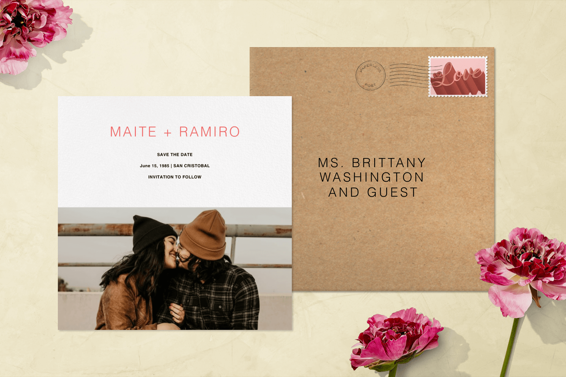 A square save the date card has a photo of a couple wearing beanies kissing on the lower half. There is a brown addressed envelope and pink flowers surrounding.