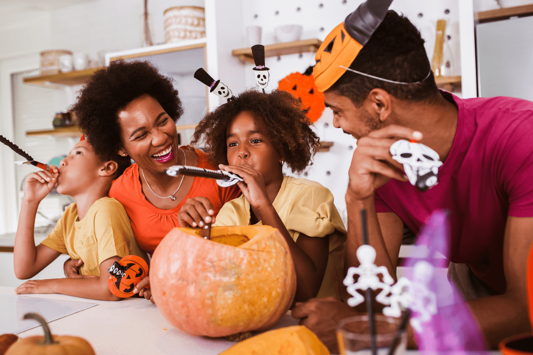 ALT: Two adults and two children wearing Halloween headbands, laughing and blowing skull party horns around a large, partly hollowed-out pumpkin at a kitchen table set with skeleton decorations, with paper jack-o'-lanterns in the background.