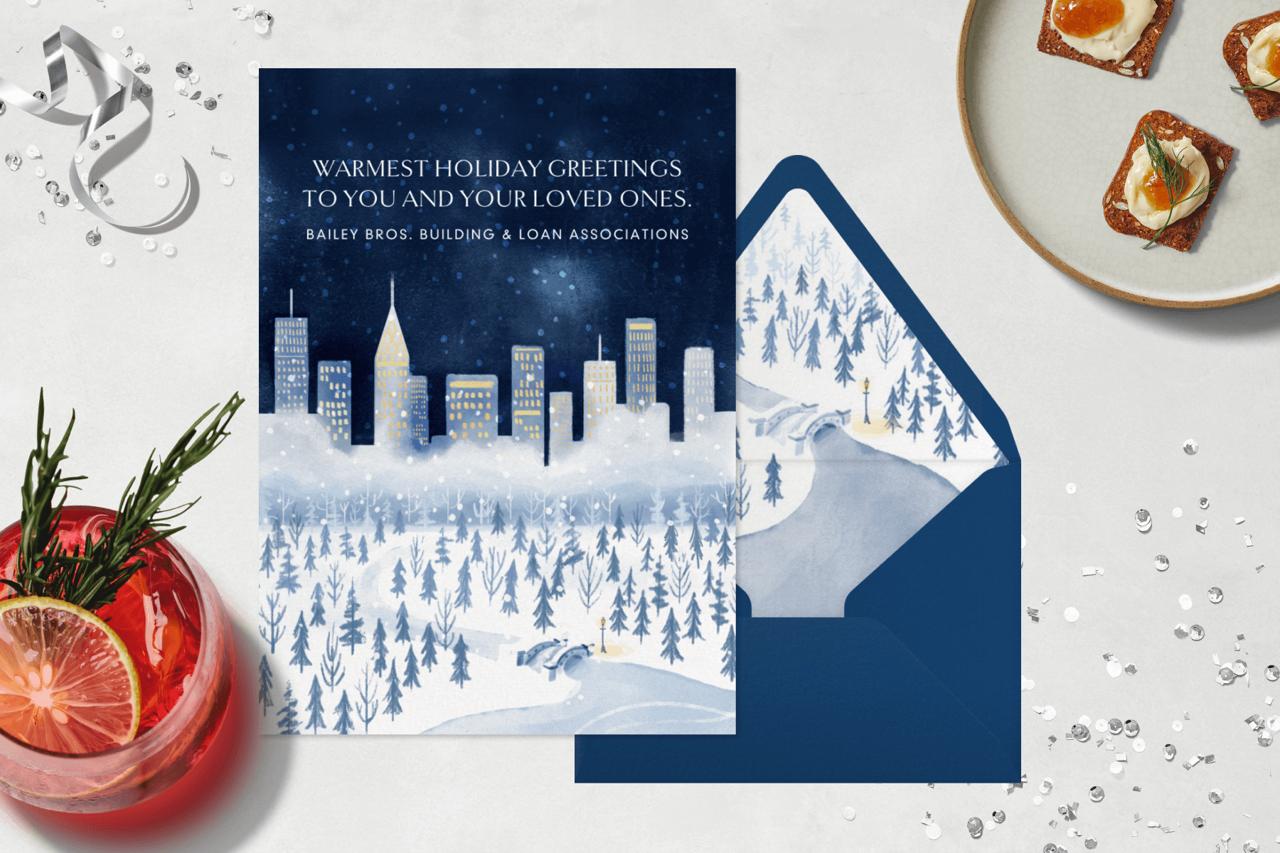 A navy blue greeting card shows a snowy winter scene beneath a city skyline at night. Nearby, a matching envelope, red cocktail, silver ribbon, confetti, and a plate with hors d'oeuvres.