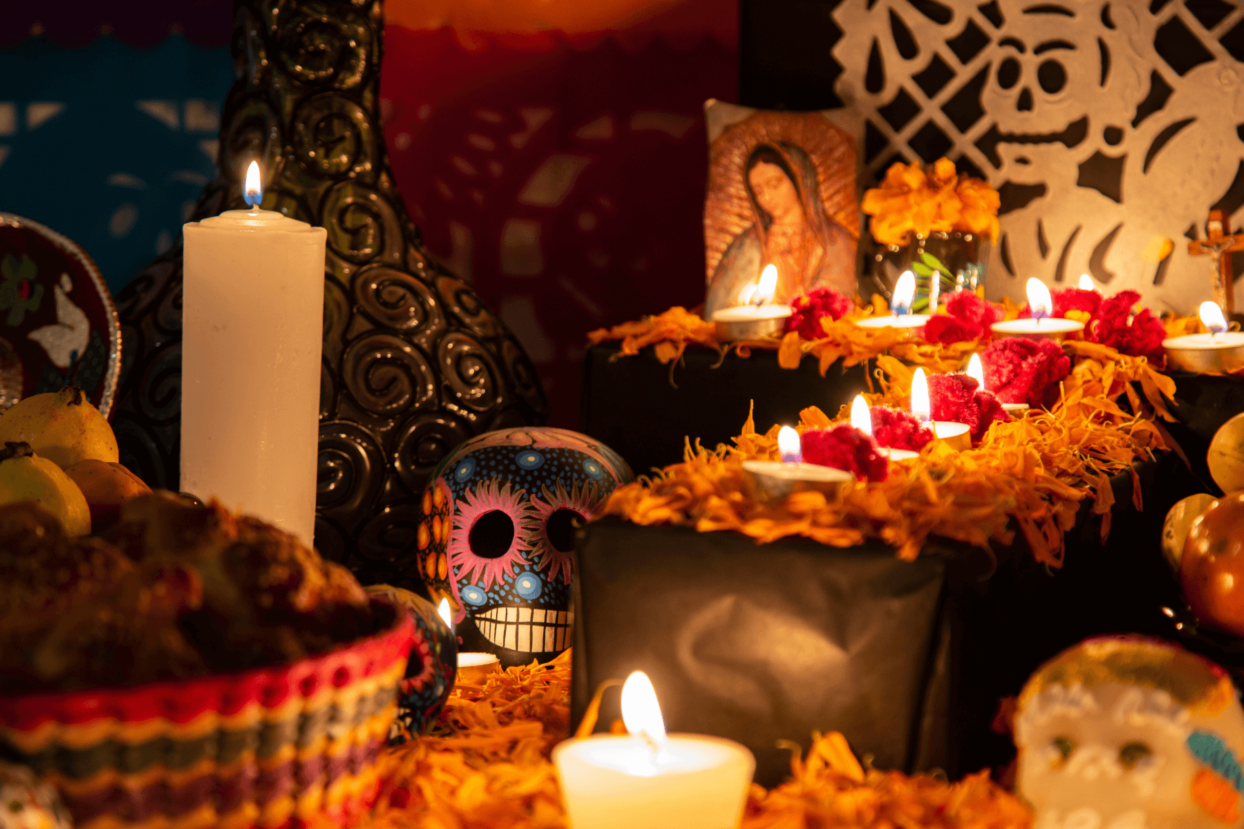 A close-up photograph of an Ofrenda decorated with candles, petals, religious imagery, and skulls.