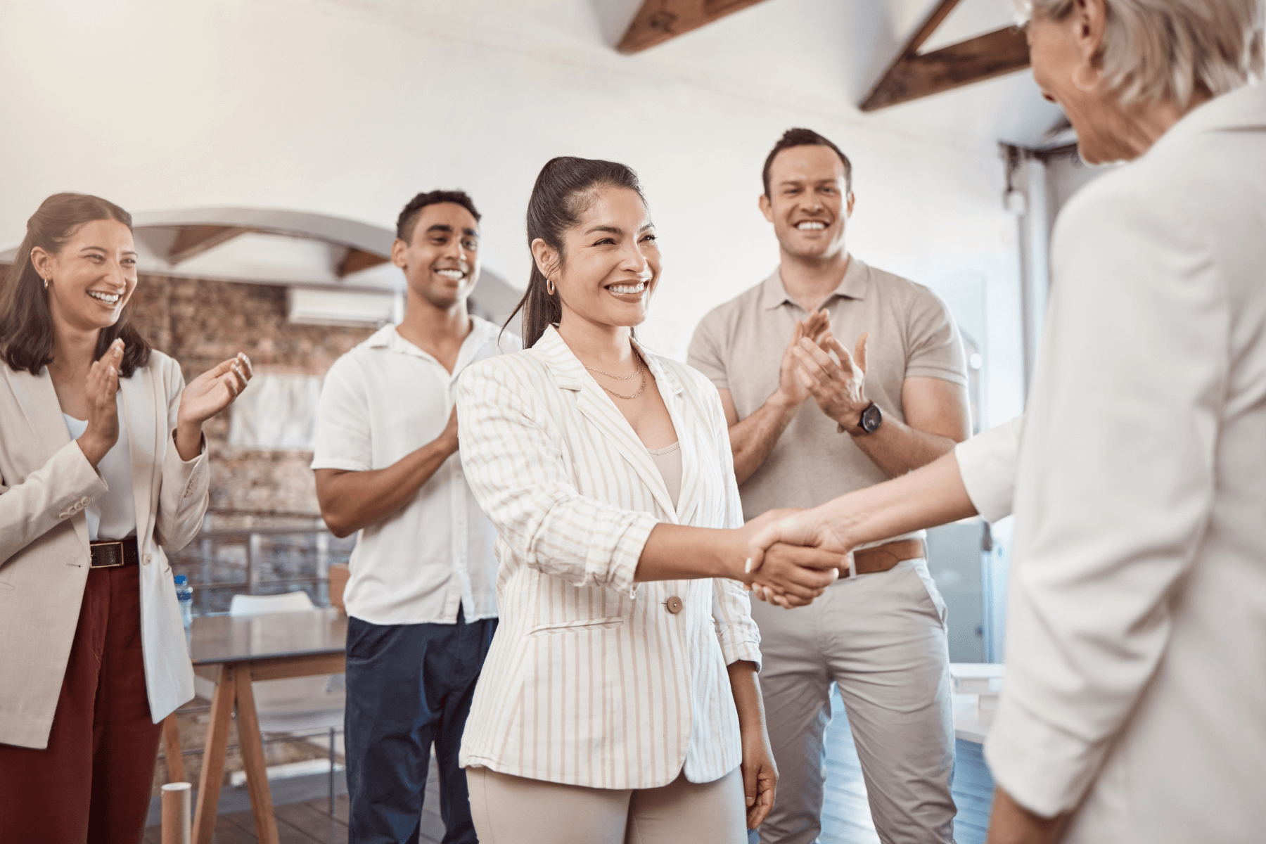 A woman in a striped suit is shaking hands with another woman in a cream colored suite while smiling widely. Two men and another woman are clapping and smiling behind her in an office setting.