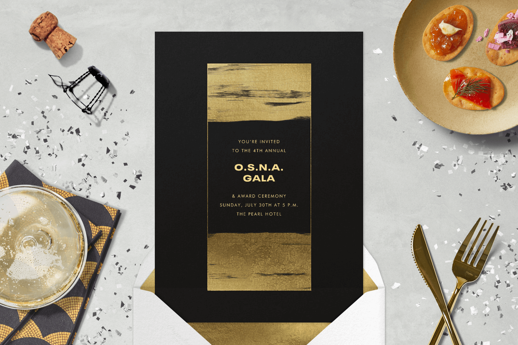 A black and gold “O.S.N.A” gala invitation with painterly gold brush strokes in the center rectangle paired with a golden envelope liner sits amongst a glass of champagne, a small plate of appetizers, a champagne cork, silver confetti, and a gold fork and knife.