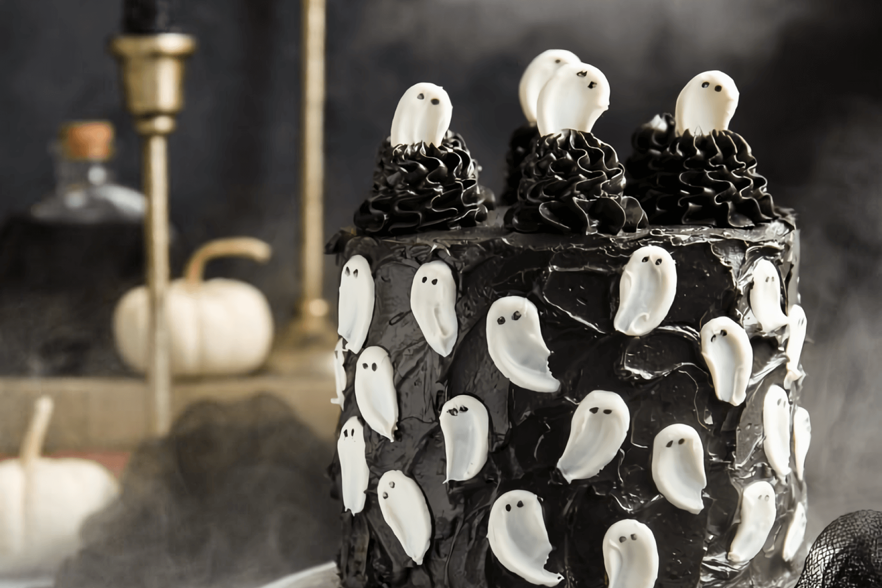 A black layer cake covered in simple white ghosts and ghosts emerging from the top, with white gourds and brass candle holders in the smoky distance.