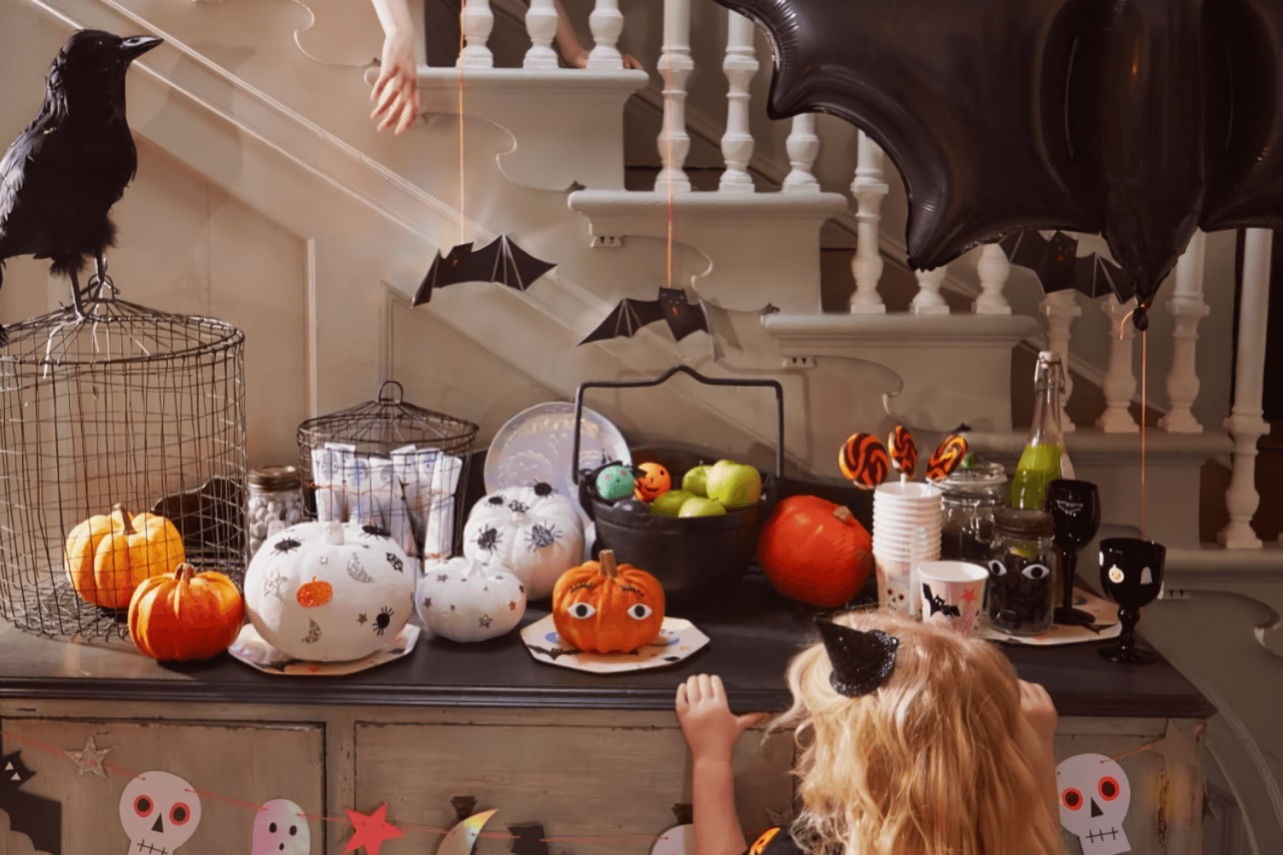 A young child wearing a small witch hat peers at a buffet table with decorated pumpkins and Halloween treats and décor in front of a white staircase.