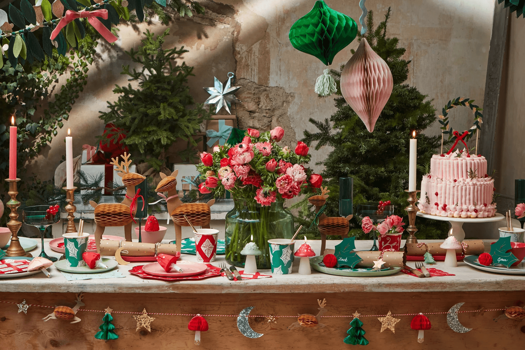 A wooden table is covered in airy Christmas décor like honeycomb items, novelty and taper candles, small evergreen trees, a pink cake, and felt holly.