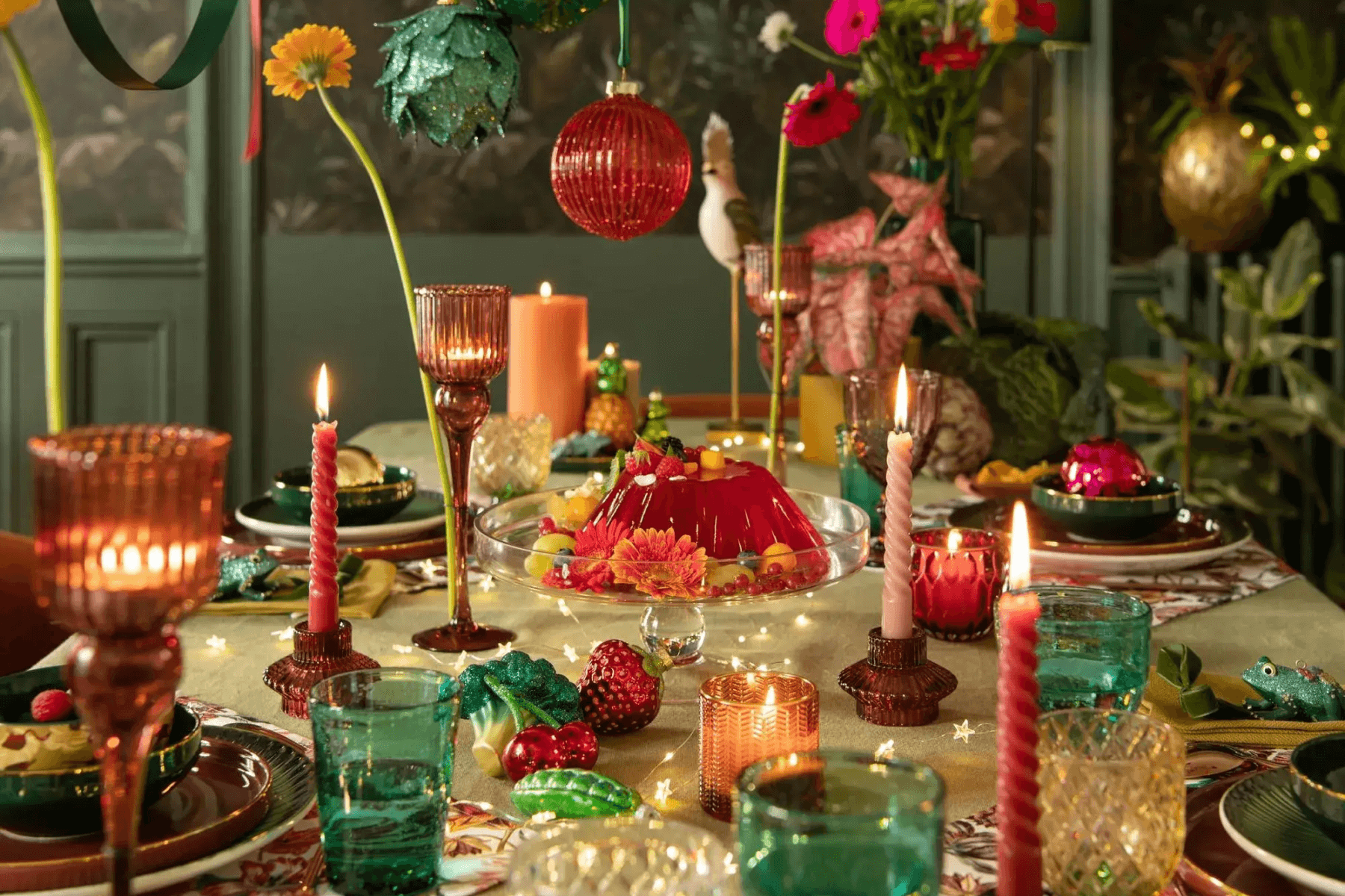A table is set with colorful glassware, food-shaped Christmas tree ornaments, taper candles, and single-stem flowers.