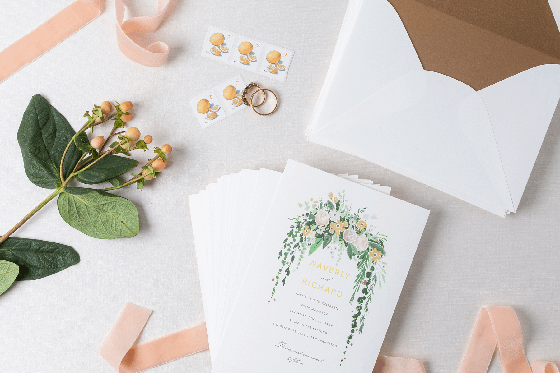 A stack of printed wedding invitations featuring a watercolor floral illustration are surrounded by pink velvet ribbon, lemon stamps, and a pair of wedding rings.