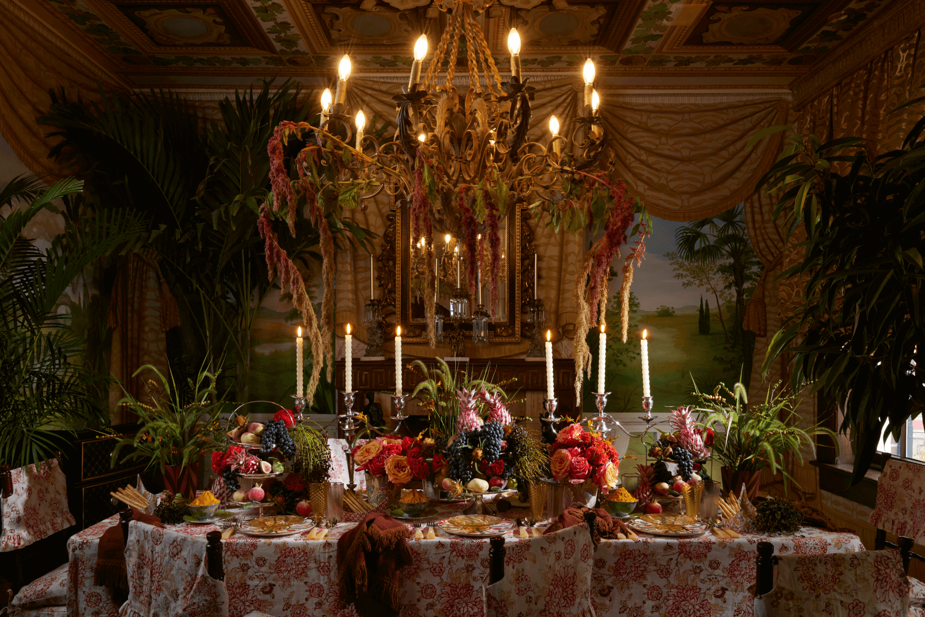 An ornate room with landscape-painted walls, gold drapes, and a large chandelier with floral arrangement is set for a meal with tall taper candles, pink toil chair covers, and lots of colorful flowers.