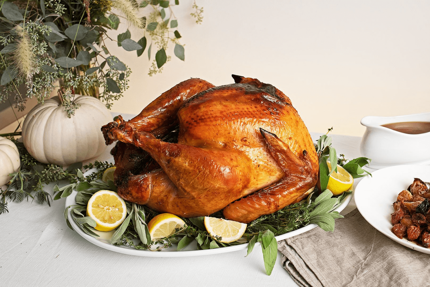 A roast turkey on a serving dish with sage leaves and lemon slices beside a white gourd and greenery arrangement.