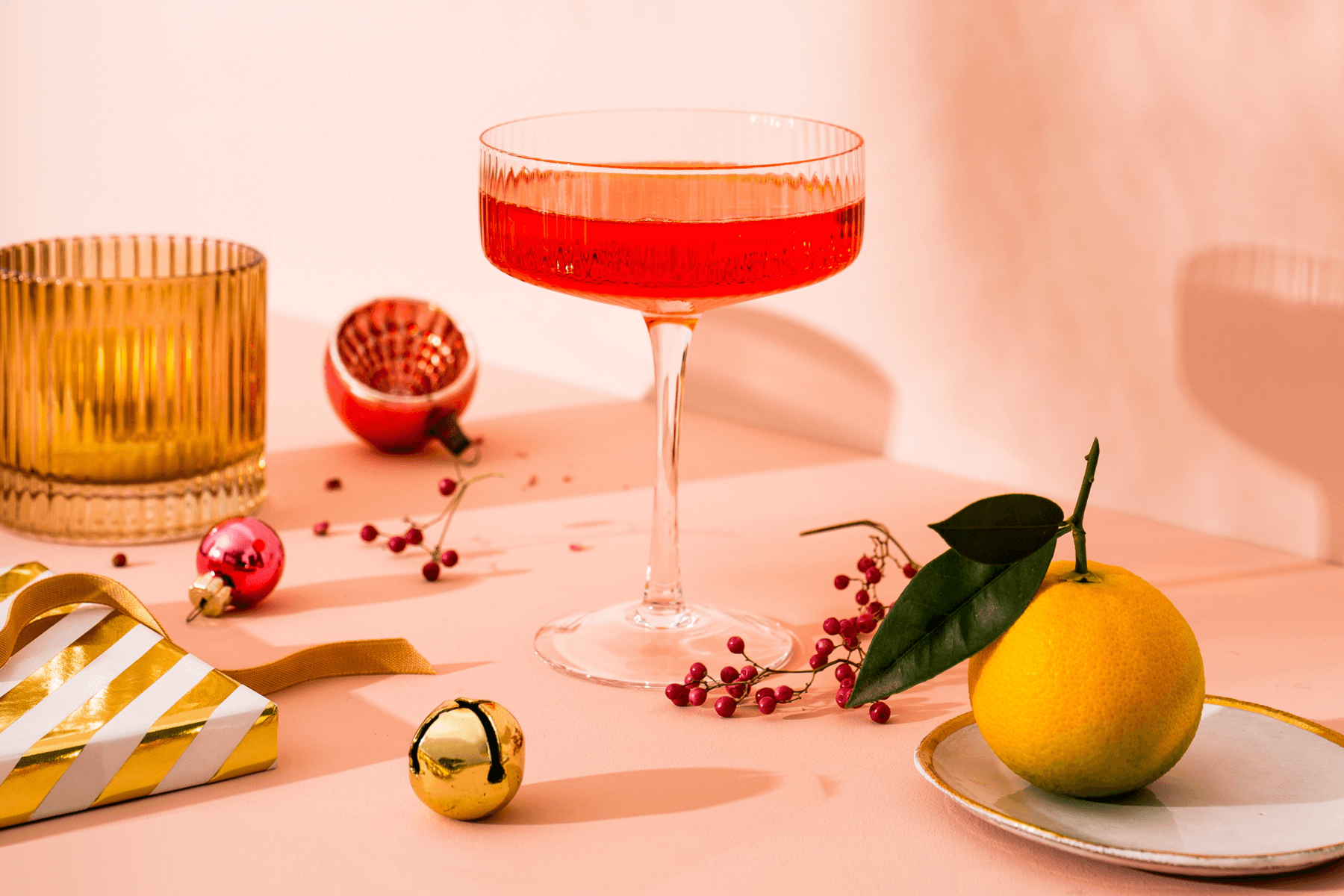 A red drink in a coupe glass surrounded by Christmas ornaments, fruit, and presents on a pink backdrop.