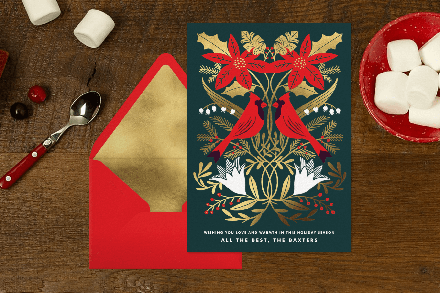 A holiday card with gold greenery, poinsettia, and cardinals beside a red envelope on a wood background with a spoon and marshmallows.