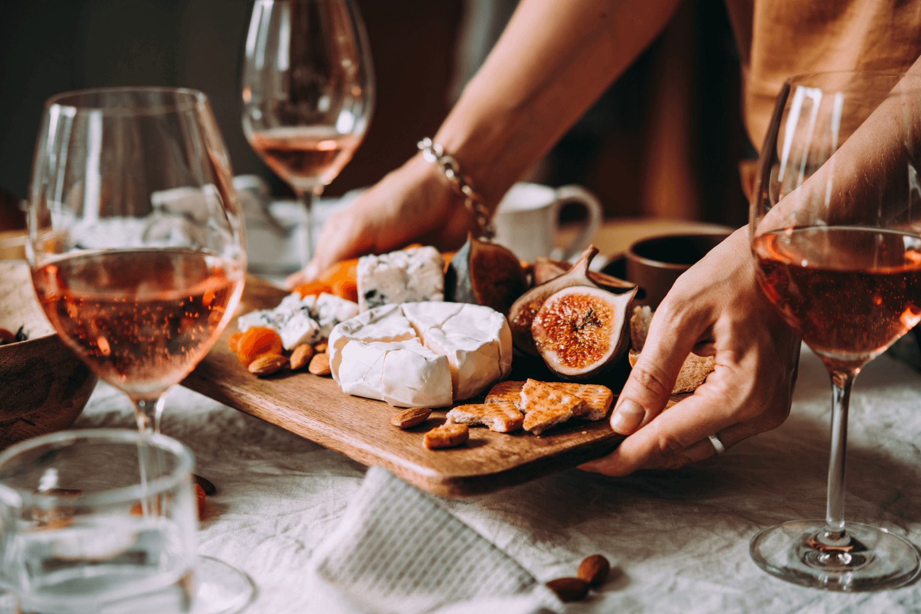 A person’s hands set down a board of cheese, figs, and nuts among wine glasses filled with rosé.