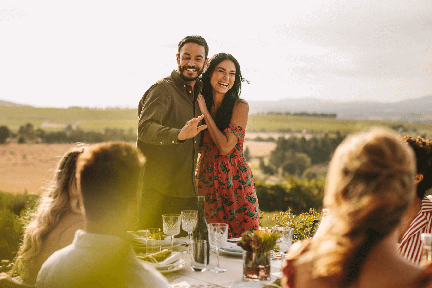 A happy couple laughs and hugs in front of a table of friends set in a rustic area.