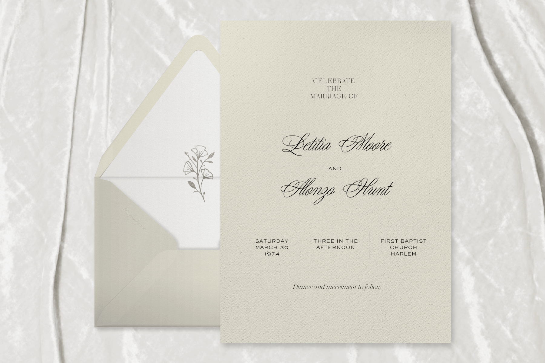 A light sage green wedding invitation on a white velvet backdrop shows the couple’s names in delicate script in the center, beside a matching envelope with a small flower bud illustration on the liner.