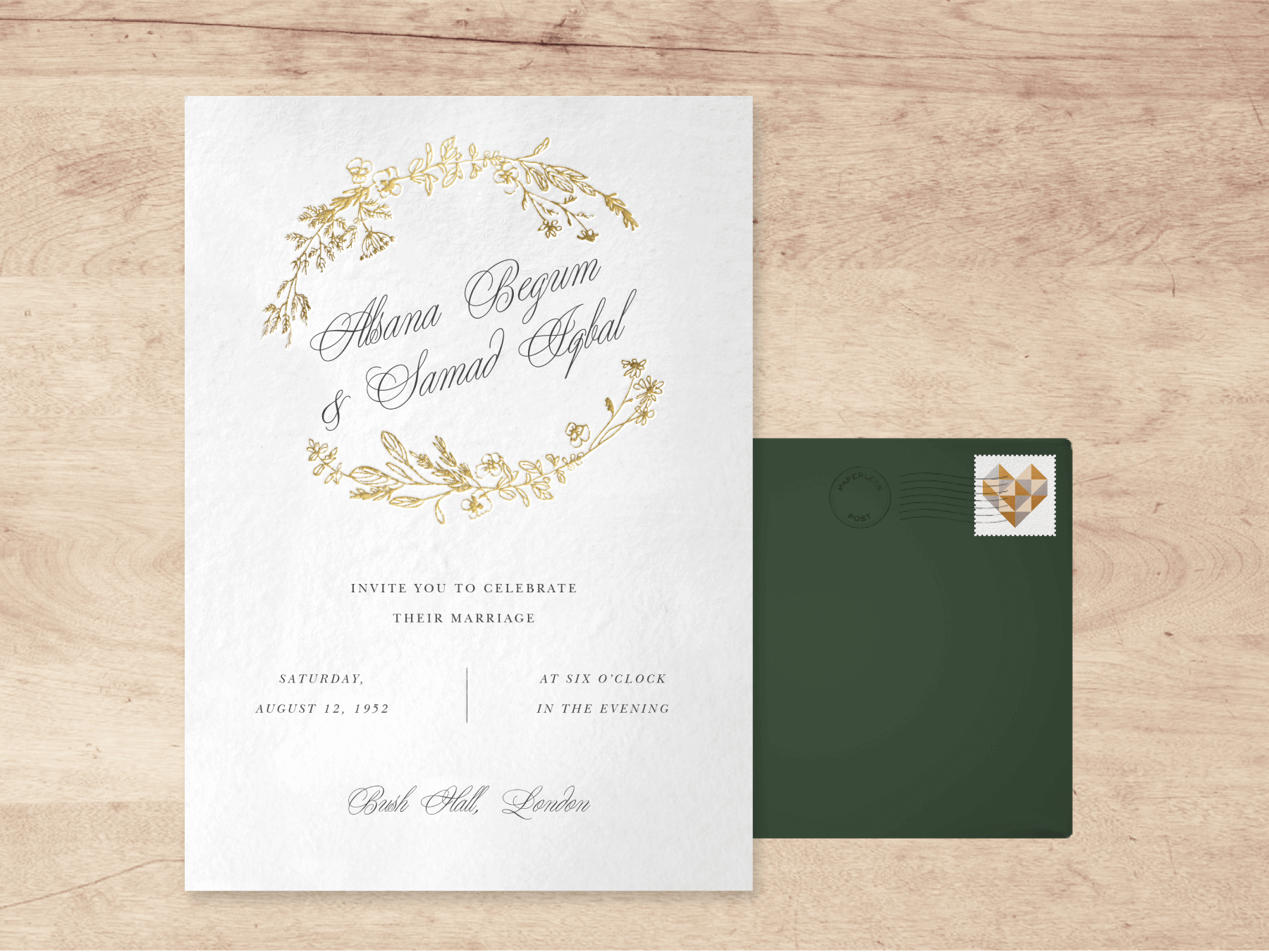 A white wedding invitation with gold sprigs circling the couple’s names written in script with a green envelope and a stamp with a geometric heart, all on a wood grain background.