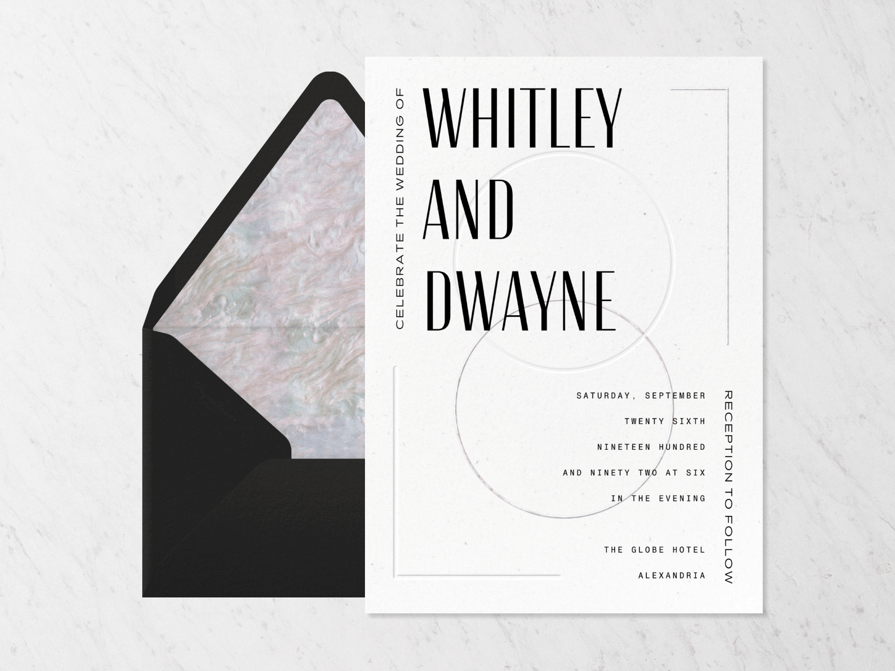 A white, minimalist wedding invitation with horizontally interlocking circles and the couple’s names written in large sans serif font beside a black envelope with marbled liner on a stone background.