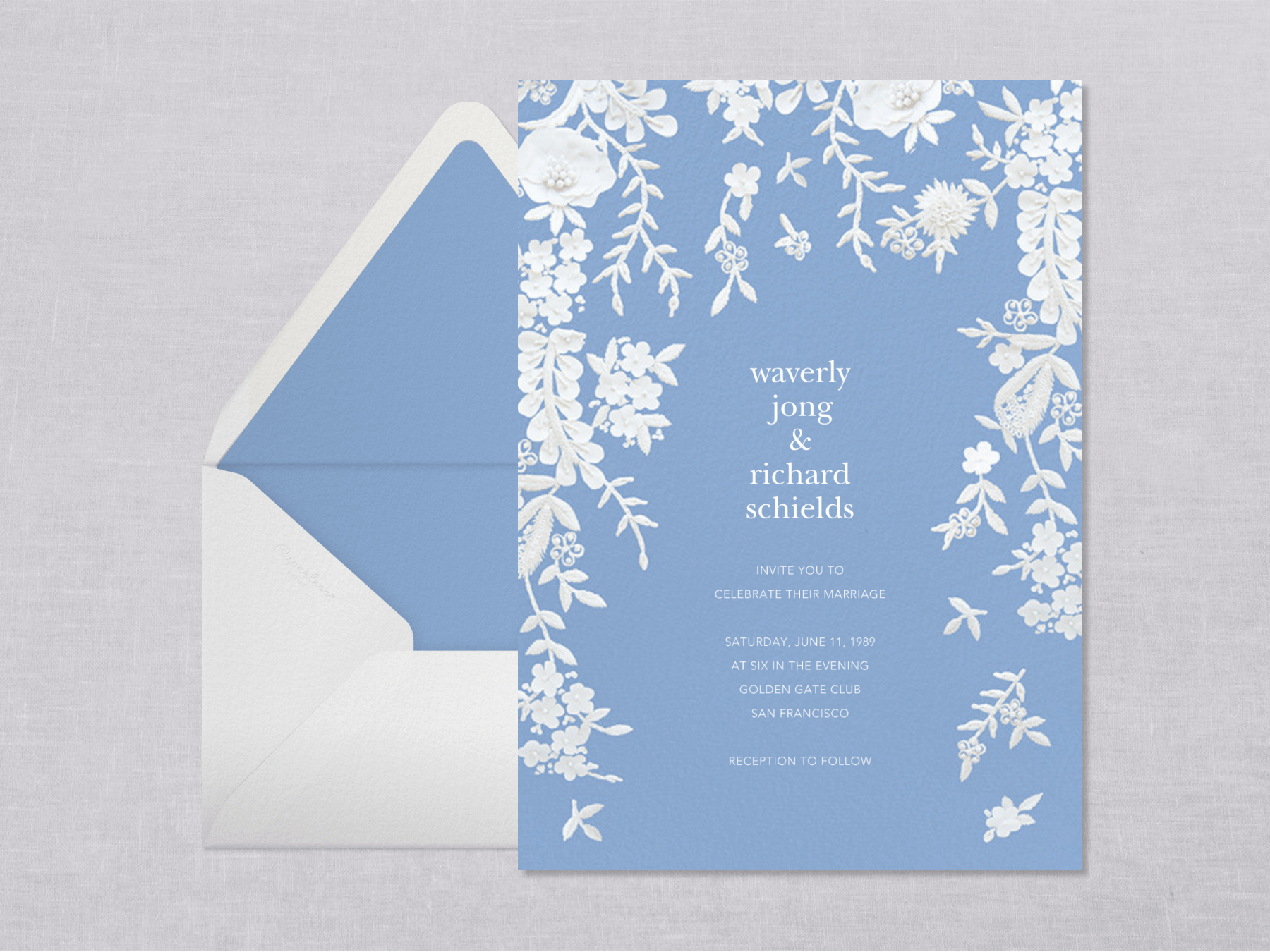 A periwinkle blue wedding invitation with white lace flowers tumbling down the edges beside a white envelope with blue liner on a gray cloth background.