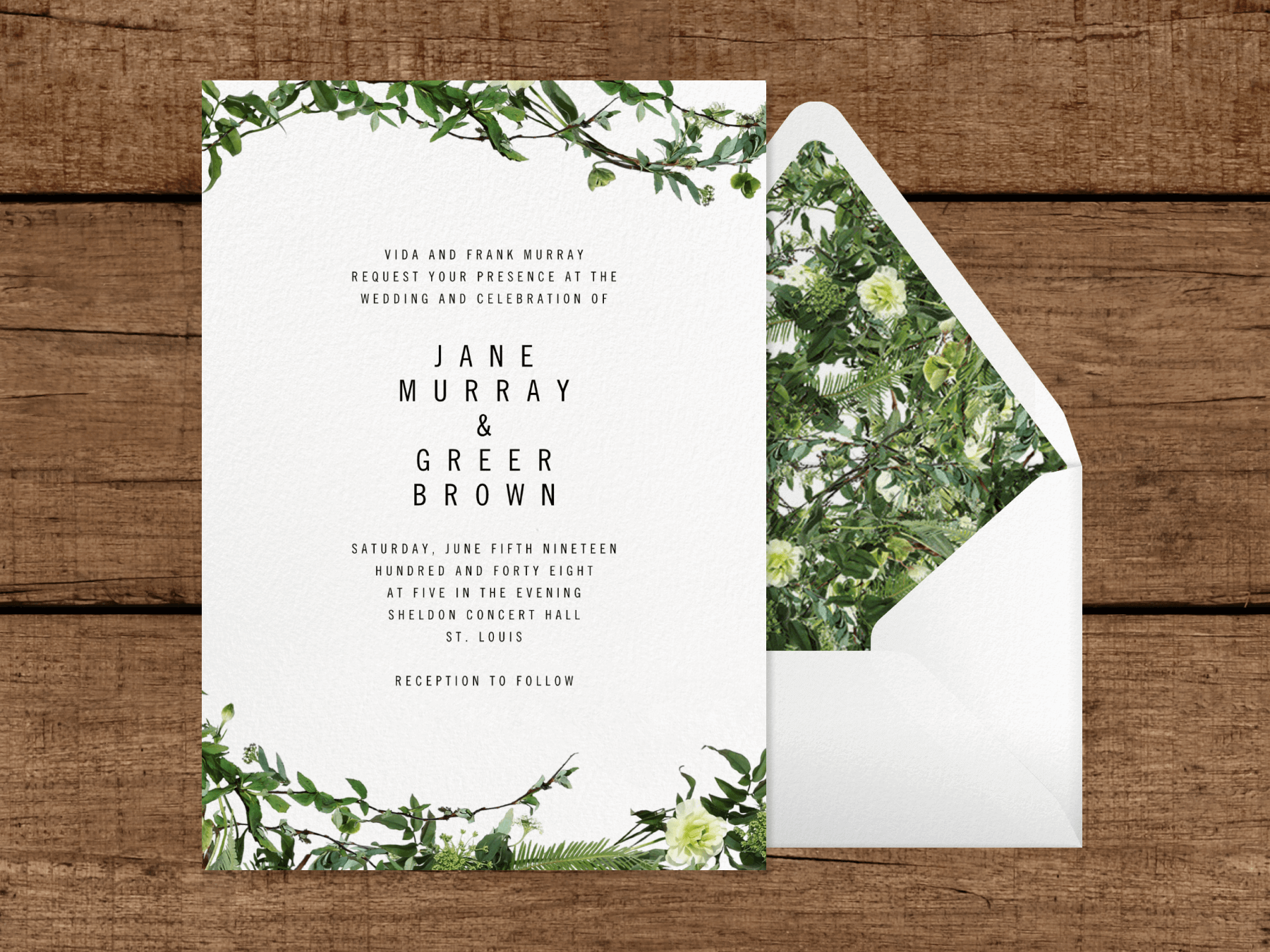 A white wedding invitation with delicate greenery on the top and bottom next to a white envelope with leafy liner, on a wood slat background.