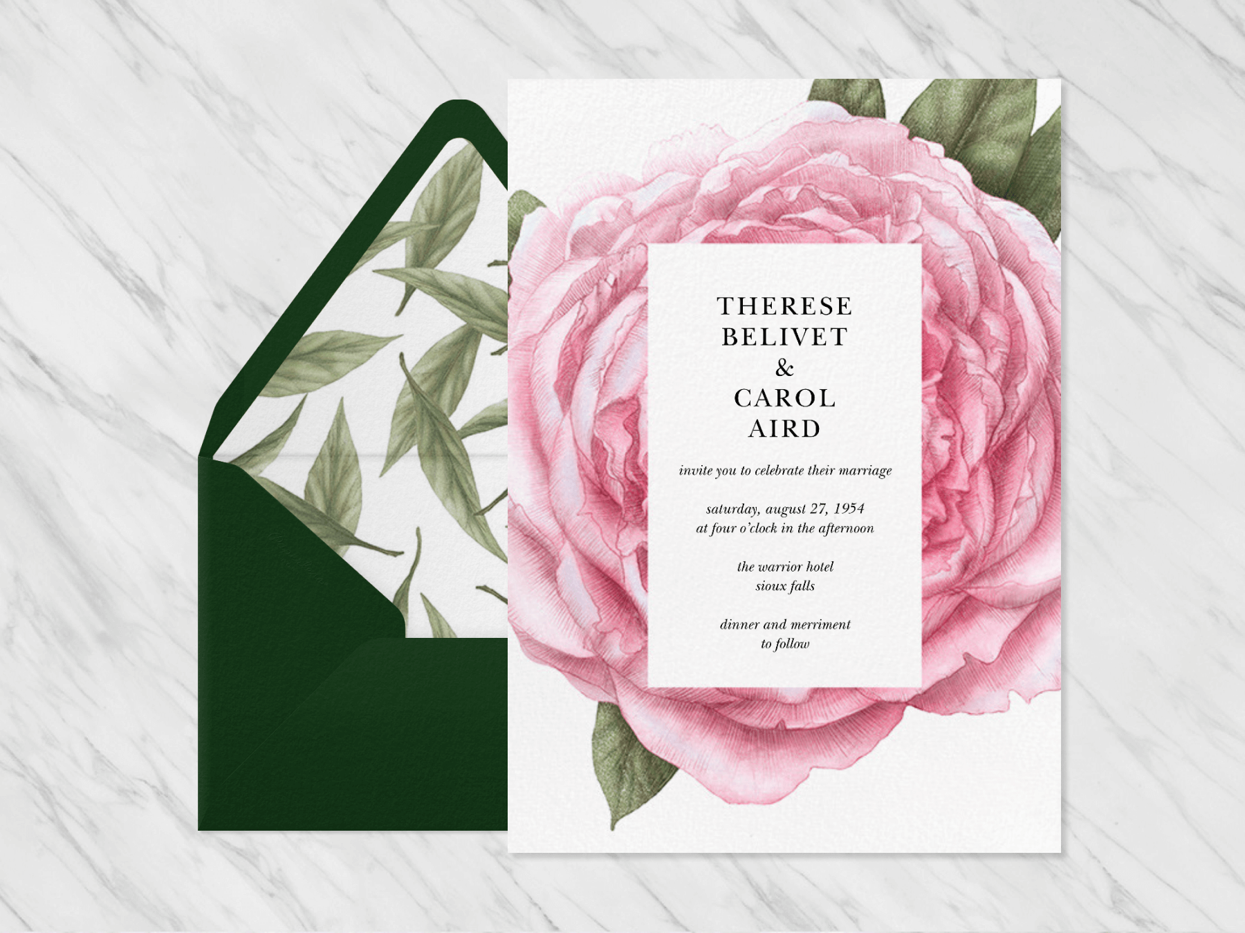 A wedding invitation with a large pink flower in the center beside a green envelope with leafy liner on a marble background.