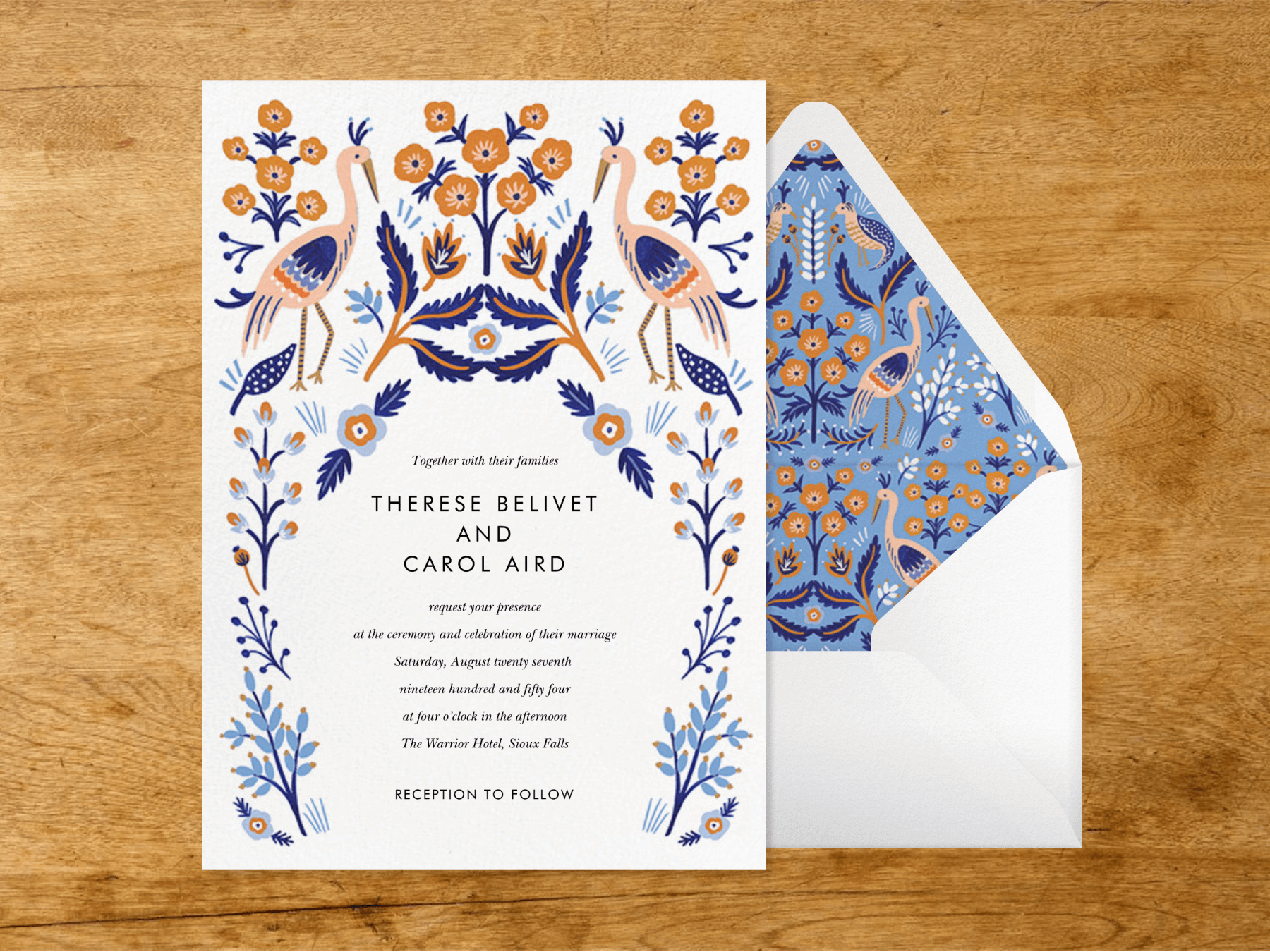 A white wedding invitation with simplistic orange and blue illustration of leaves, flowers, and long-necked birds forming a border beside a white envelope with a matching illustrated liner on a wood grain background.