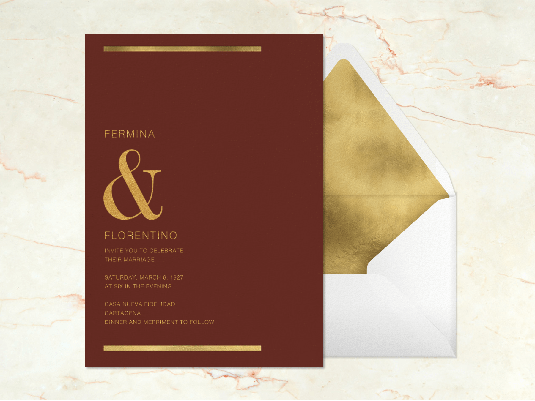 A burgundy wedding invitation with gold lines at the top and bottom and a large gold ampersand between the couple’s names beside a white envelope with gold liner on a pink marbled background.