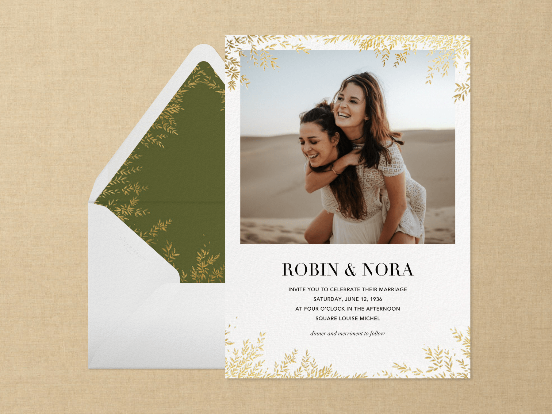 A white wedding invitation with a photo of a smiling couple piggybacking in the desert with gold leaves creeping in on the top and bottom beside a white envelope with green and gold leaf liner on a neutral textured background.