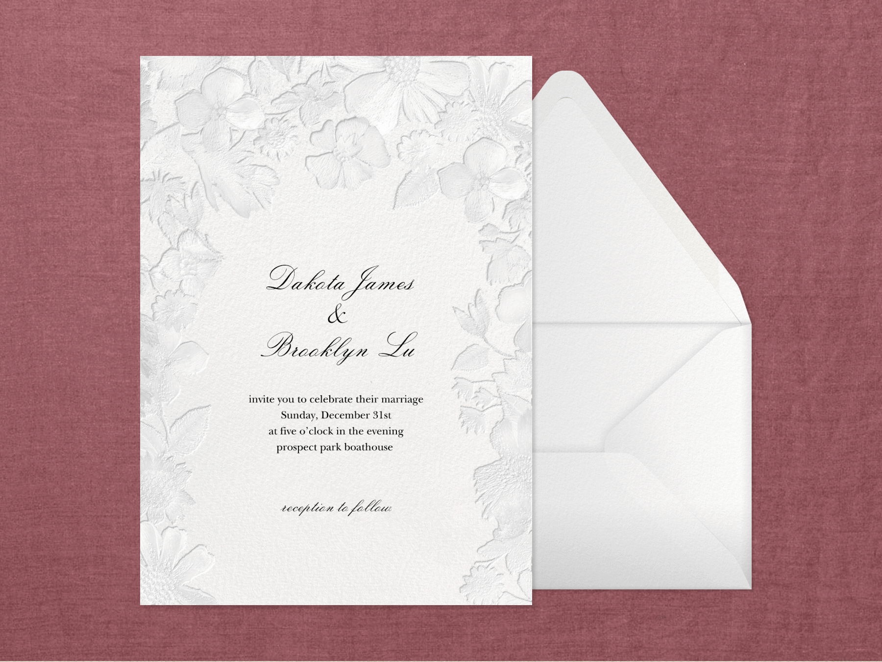 A wedding invitation with blind embossed flowers around the border next to a white envelope on a textured oxblood background.