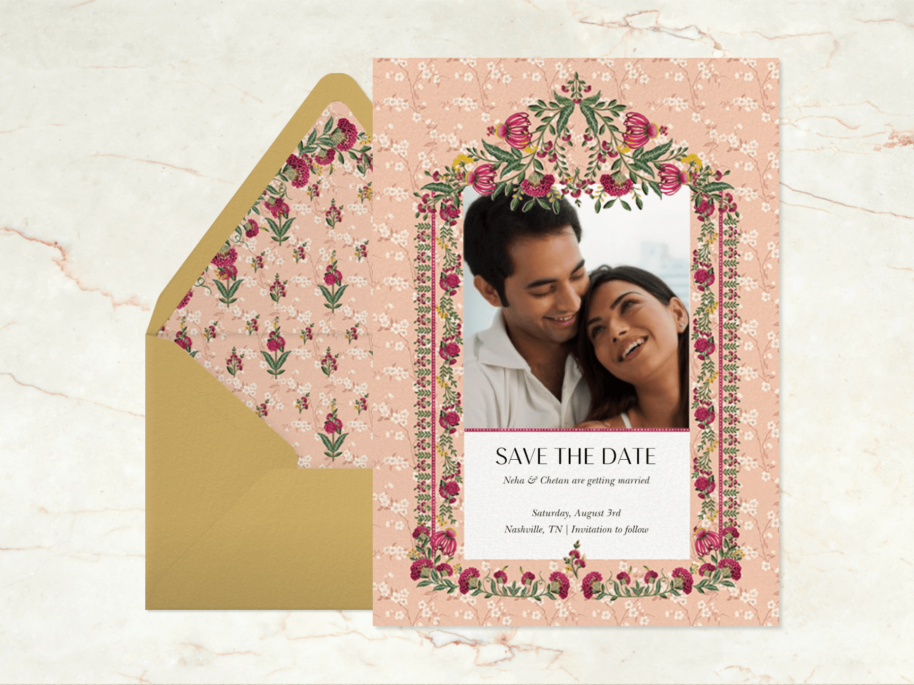 A light pink wedding invitation with a photo of a couple surrounded by a fuchsia, green, and yellow floral border next to a gold envelope with matching liner on a marble background.