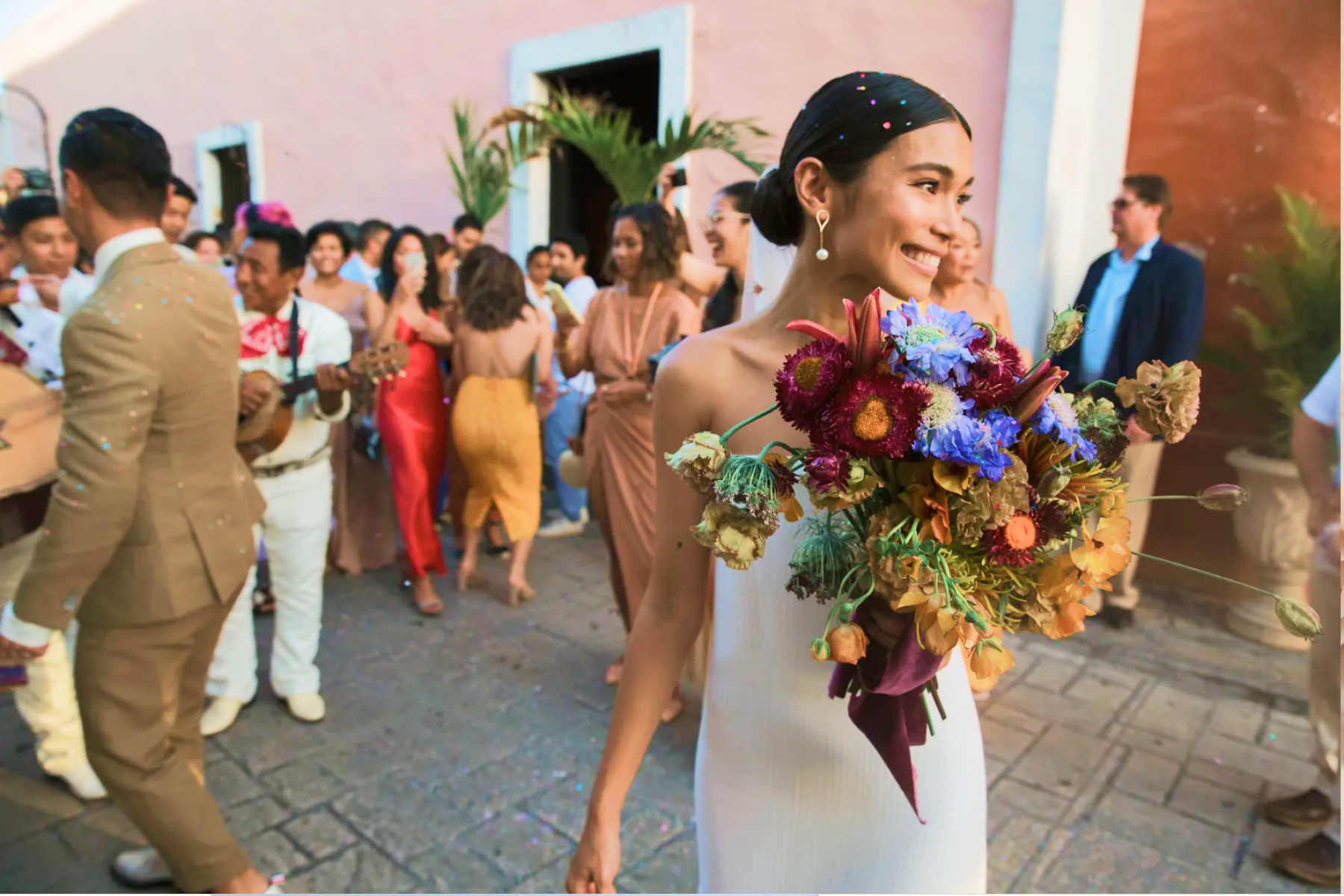 Photograph of a bride surrounded by loved ones at her wedding. She is holding a bouquet and smiling towards something off-camera.